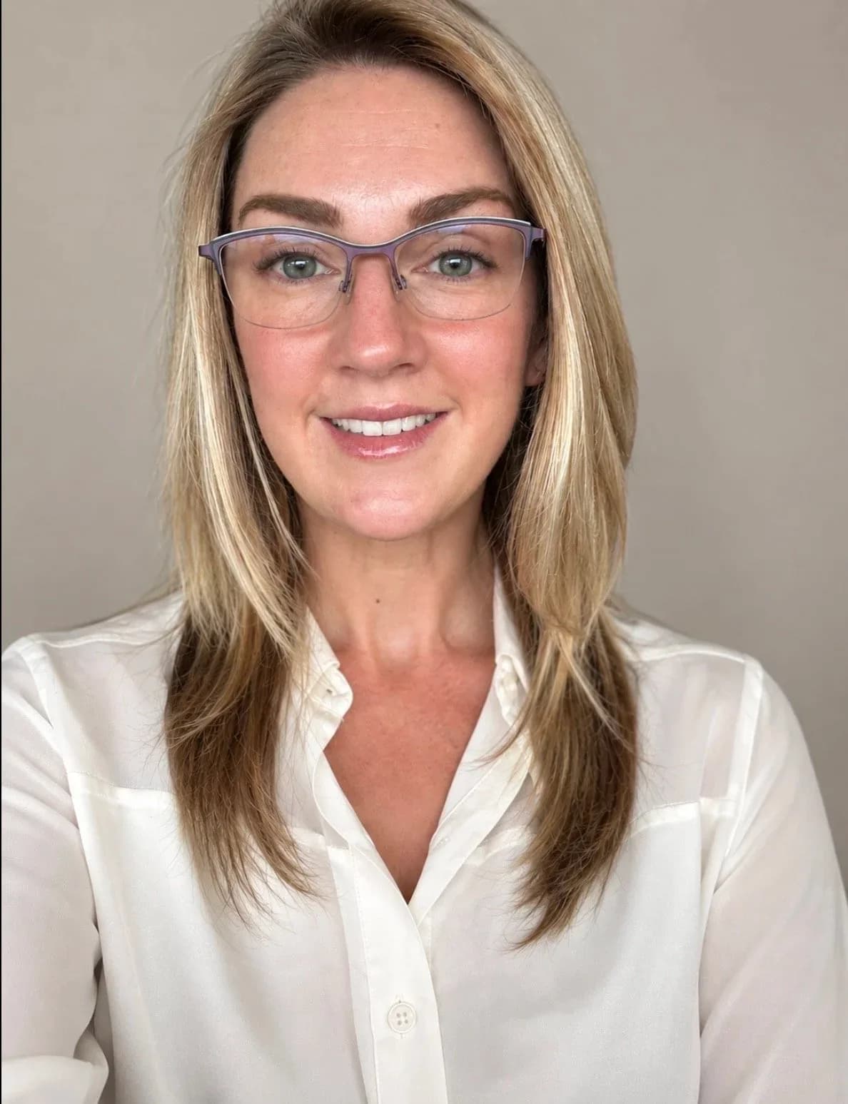 Angel Wallace, with shoulder-length blonde hair, glasses and a white button-up shirt, smiling at the camera against a plain beige background