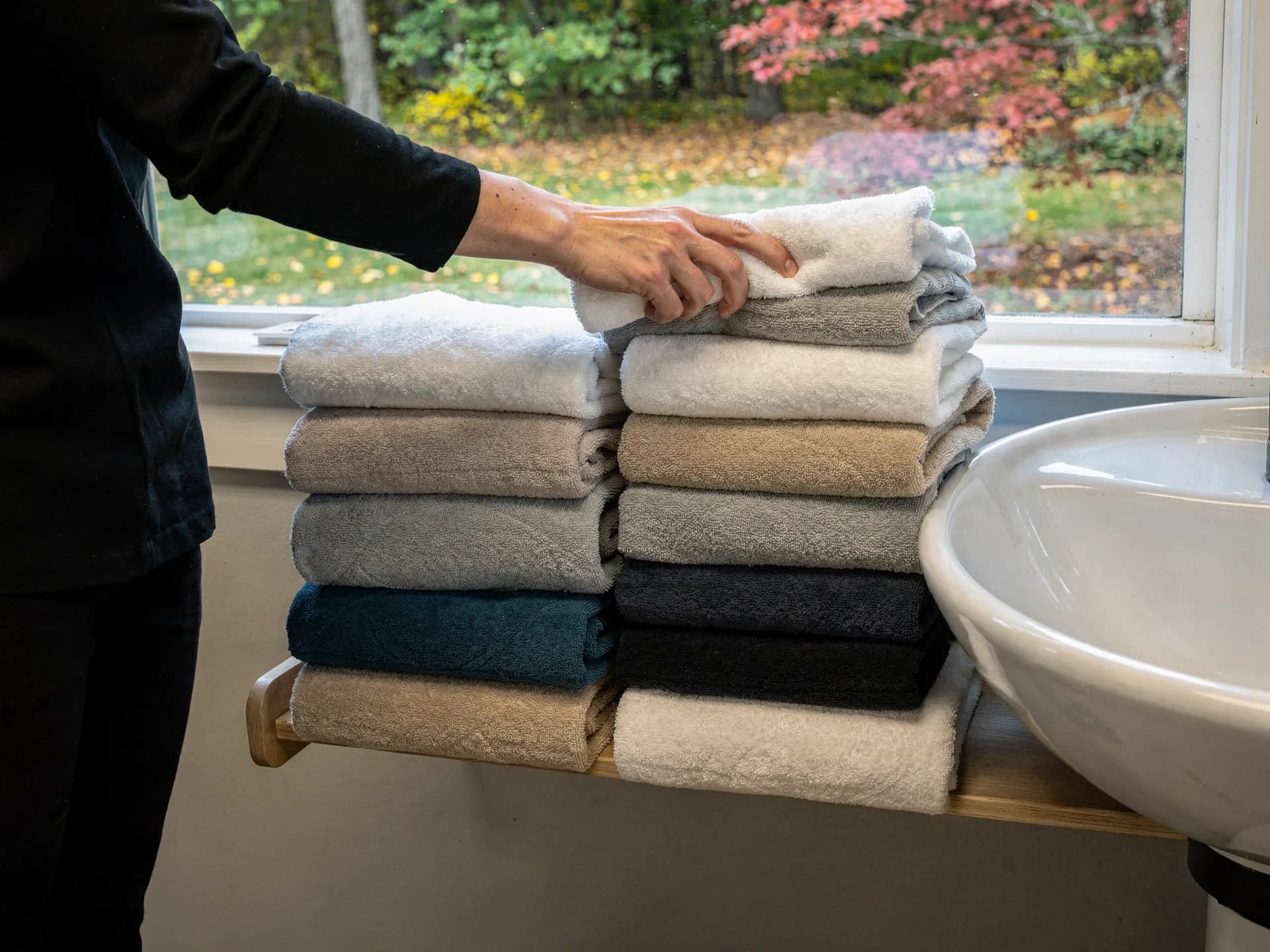A hand in a black sleeve sets a folded towel on stacks of clean salon towels beside a basin.