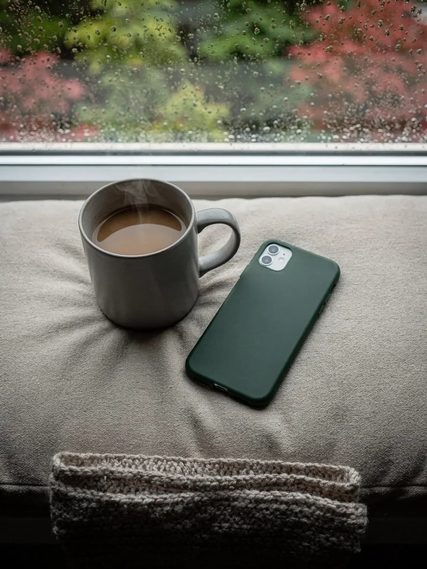 A steaming mug and a face-down phone rest on a window seat while rain beads on the glass.