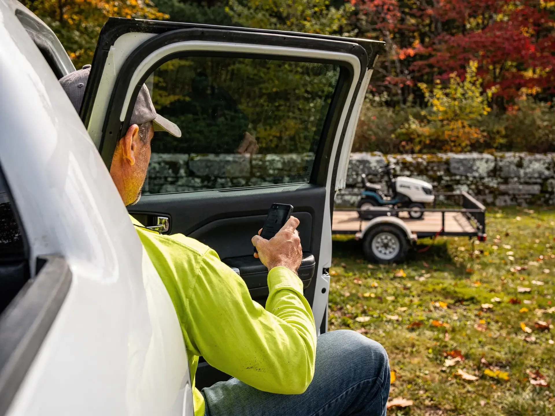 A lawn owner in hi-vis sits in his open truck door holding a phone, facing a trailer with a ride-on mower.