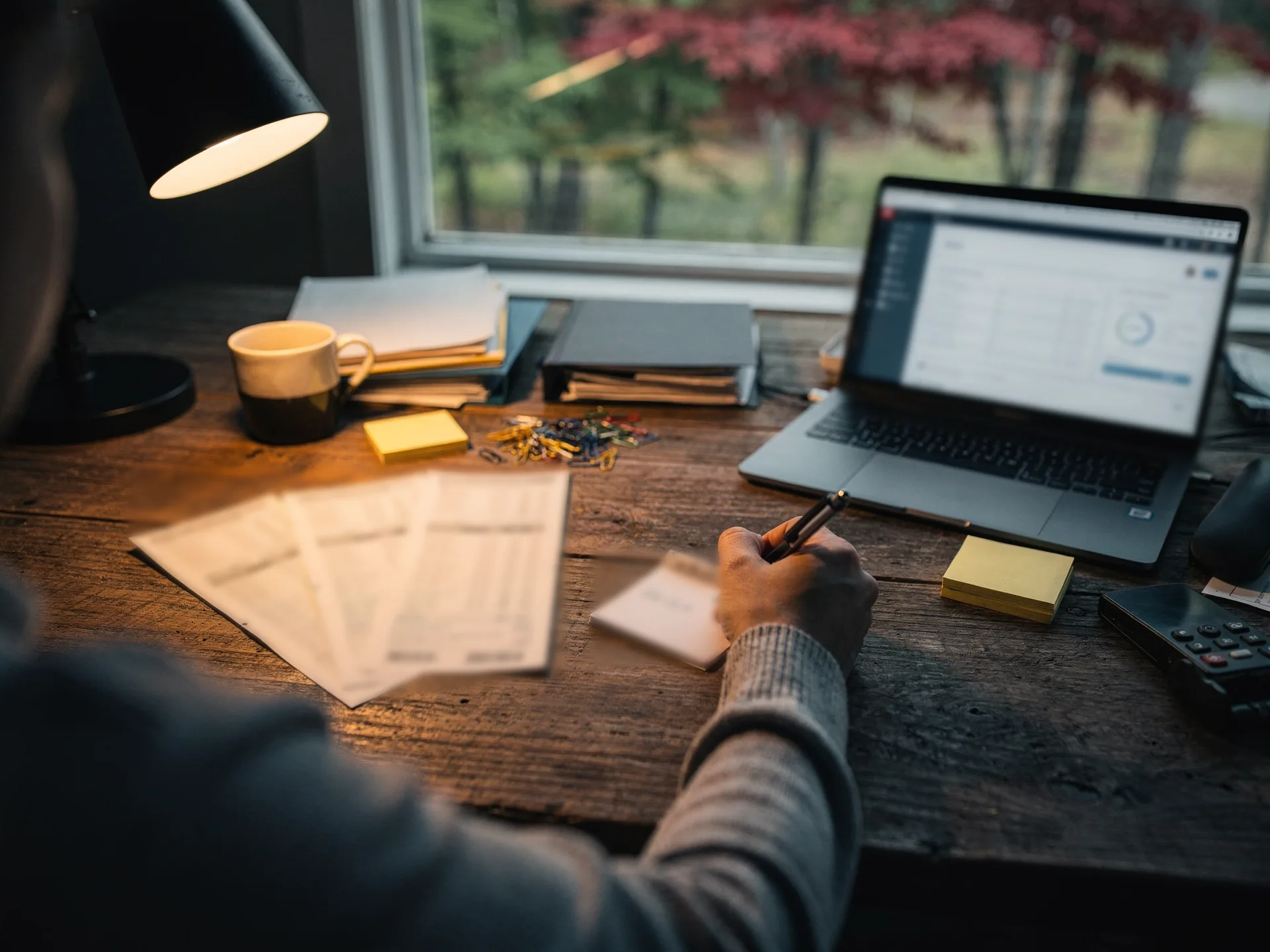 Over a shoulder in a grey knit sleeve, a hand jots figures on a small notepad beside three fanned bank statements, a laptop, a mug and a desk lamp on a worn wooden desk at dusk.