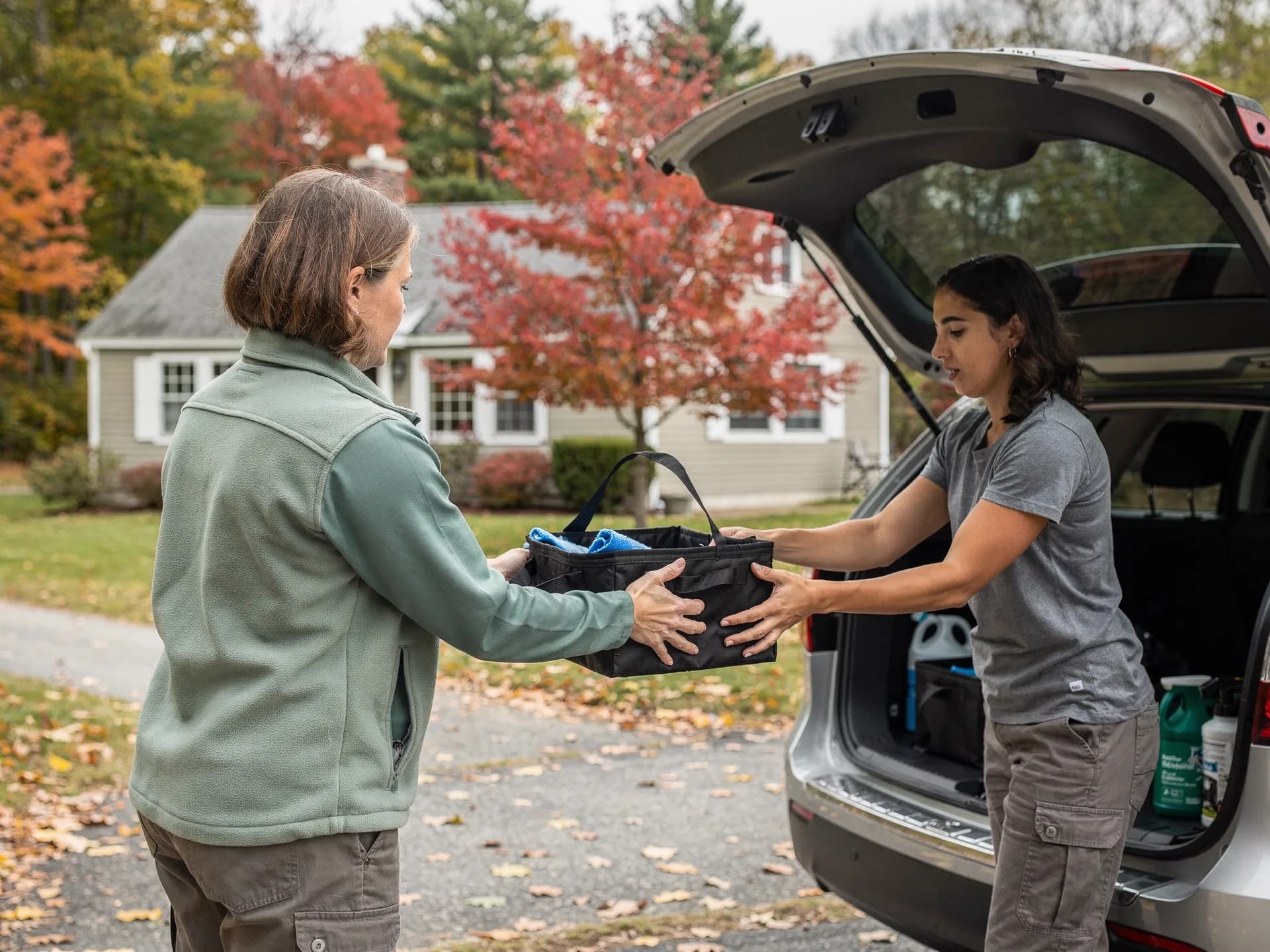 A cleaning-company owner in a sage fleece hands a black cleaning caddy to her helper at the open hatch of a car in a client's driveway.