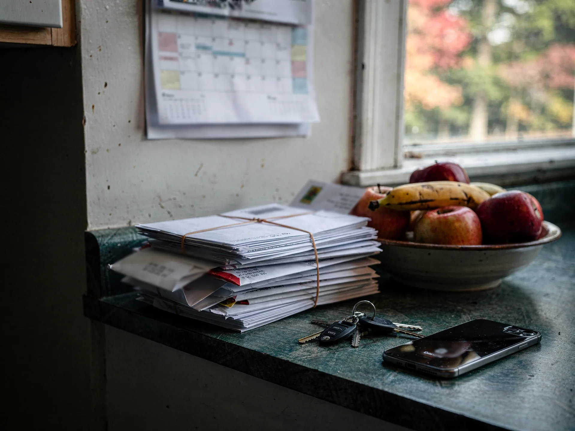 A thick bundle of unopened envelopes tied with string sits on a kitchen counter beside keys, a phone and a bowl of apples and bananas, a wall calendar hanging above and autumn trees out the window.