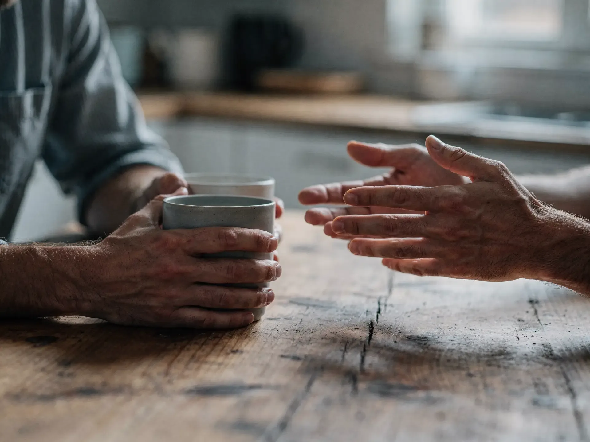 Across a wooden kitchen table, one person holds a mug while the other gestures mid-explanation.