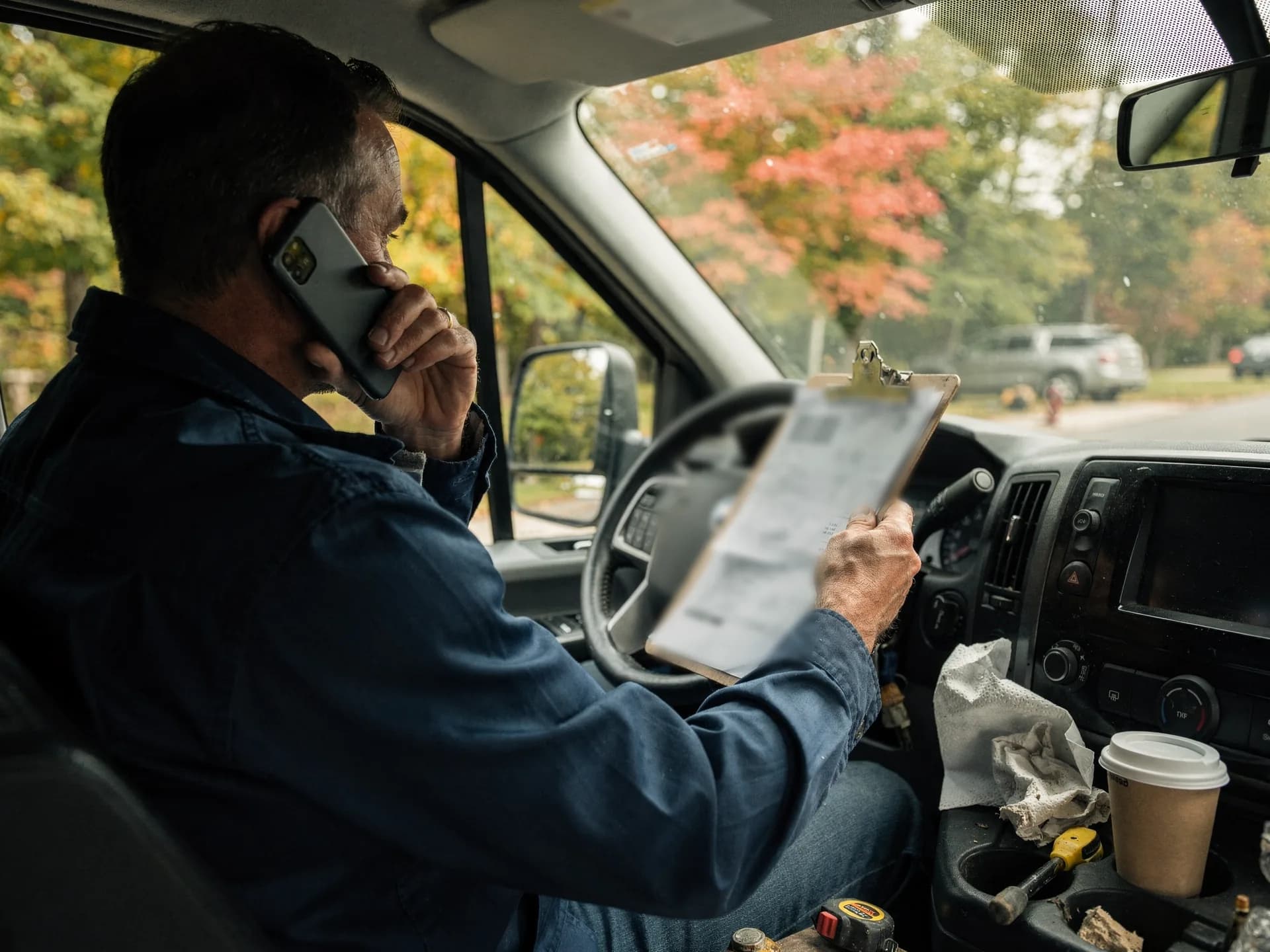 A man in a denim work shirt sits in the driver's seat of his van on a phone call, holding up a clipboard with a printed statement, a coffee cup and a tape measure in the console.