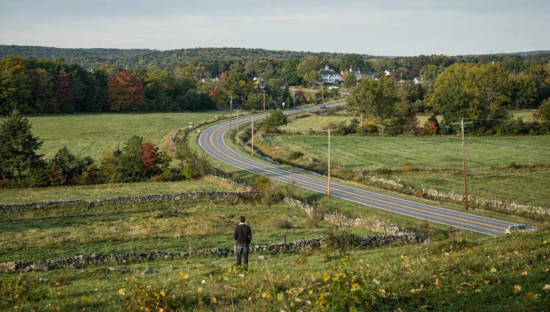 From a hillside, a man looks down a two-lane road winding past fields and stone walls toward a white village.