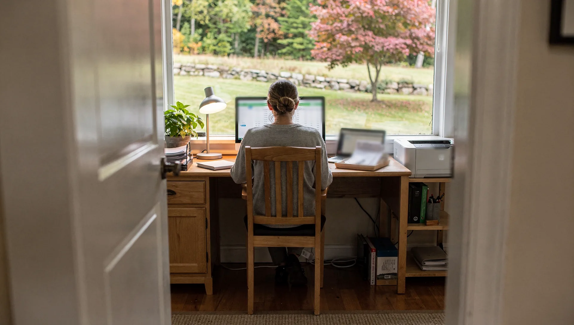 Seen through an open door, a woman with her hair in a bun sits with her back to the camera at a wooden desk, working on a monitor of spreadsheet rows beside a laptop, a printer and a tray of papers, a lawn and stone wall outside the window.
