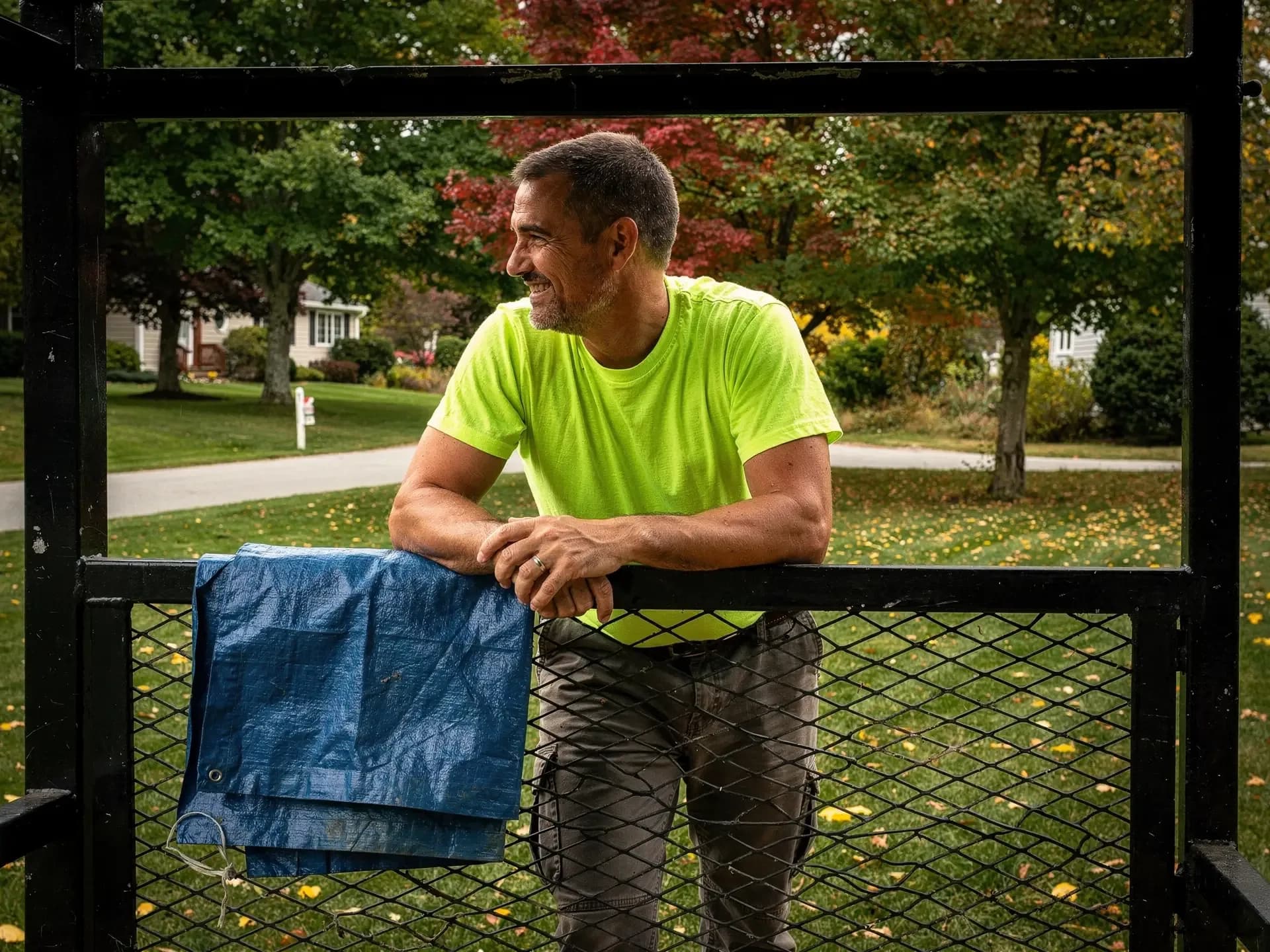 A smiling lawn-care owner in a hi-vis tee leans on the gate of his equipment trailer beside a suburban lawn and red maple.