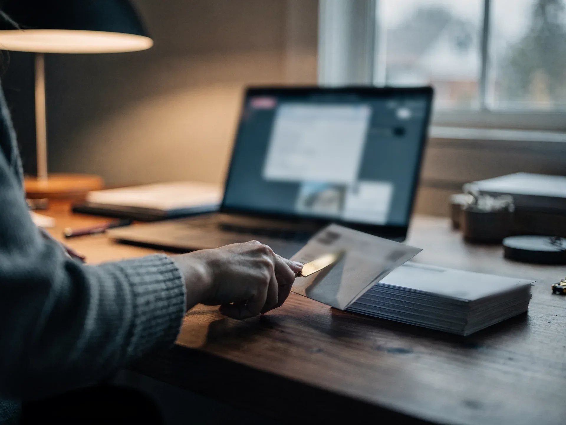 A hand in a grey knit sleeve slits open a bank statement envelope with a brass letter opener beside a stack of opened statements.