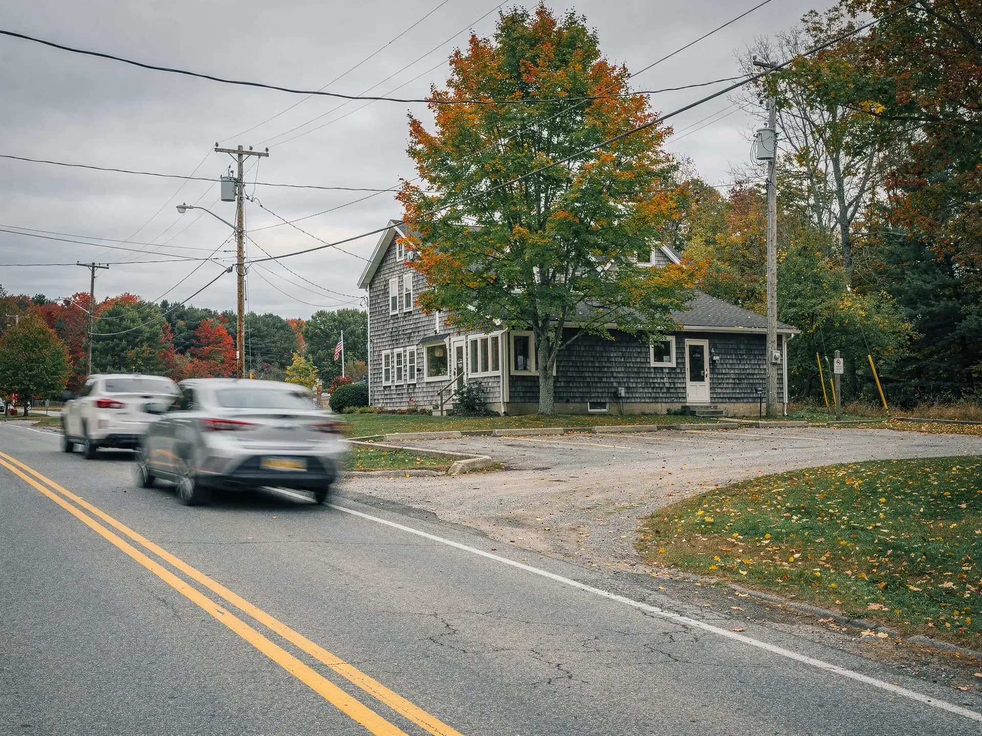 A gray-shingled small office building with a gravel lot on a two-lane road, cars blurred as they pass a turning maple.