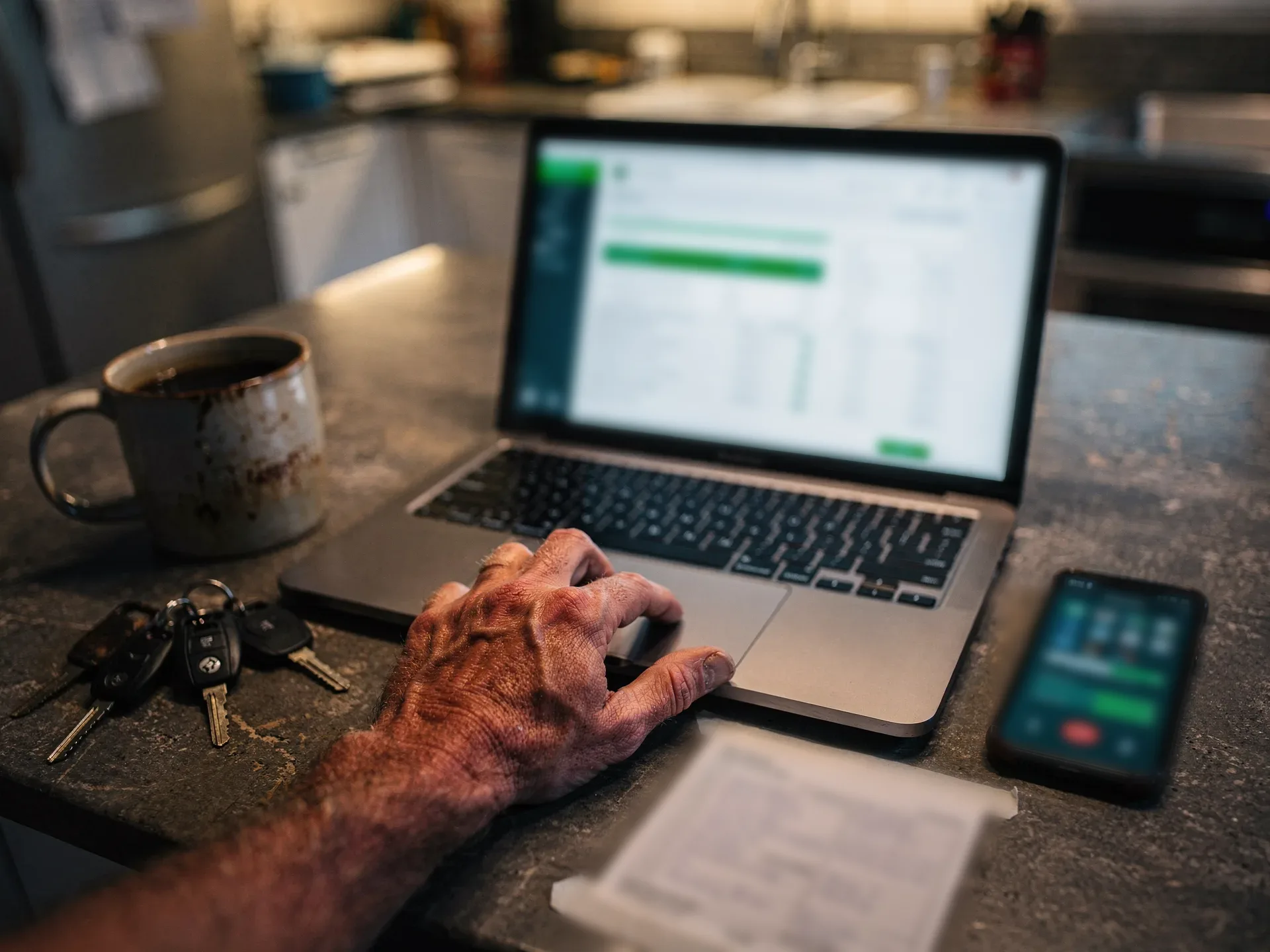 A weathered hand rests on a laptop trackpad showing an accounting screen with green accents, a phone on a call beside it and truck keys by a mug.