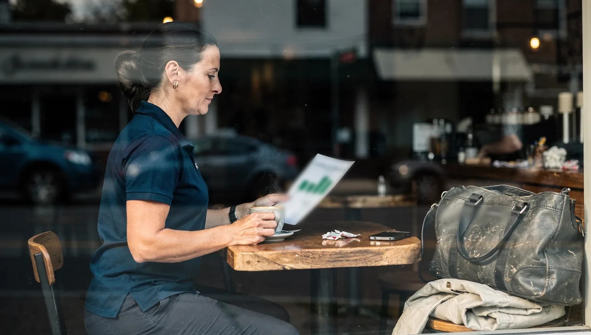 Through a cafe window, a woman in a navy work polo sits at the counter with a latte, reading a one-page report with a small green chart, her tote and jacket on the next stool.