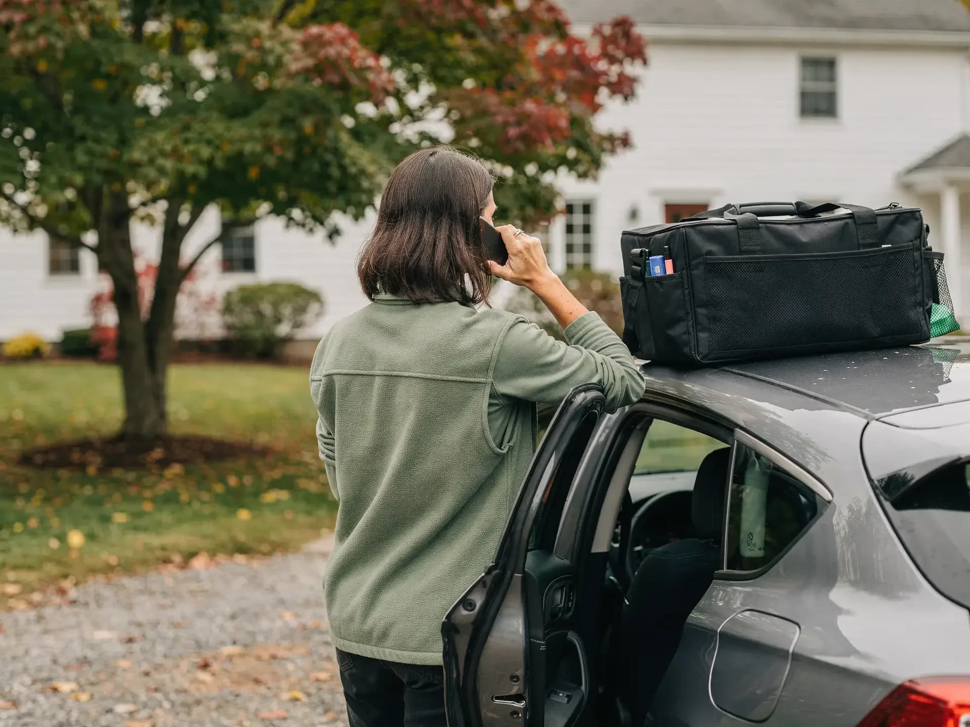 A woman in a fleece seen from behind talks on her phone while leaning on her open car door in a client's driveway.