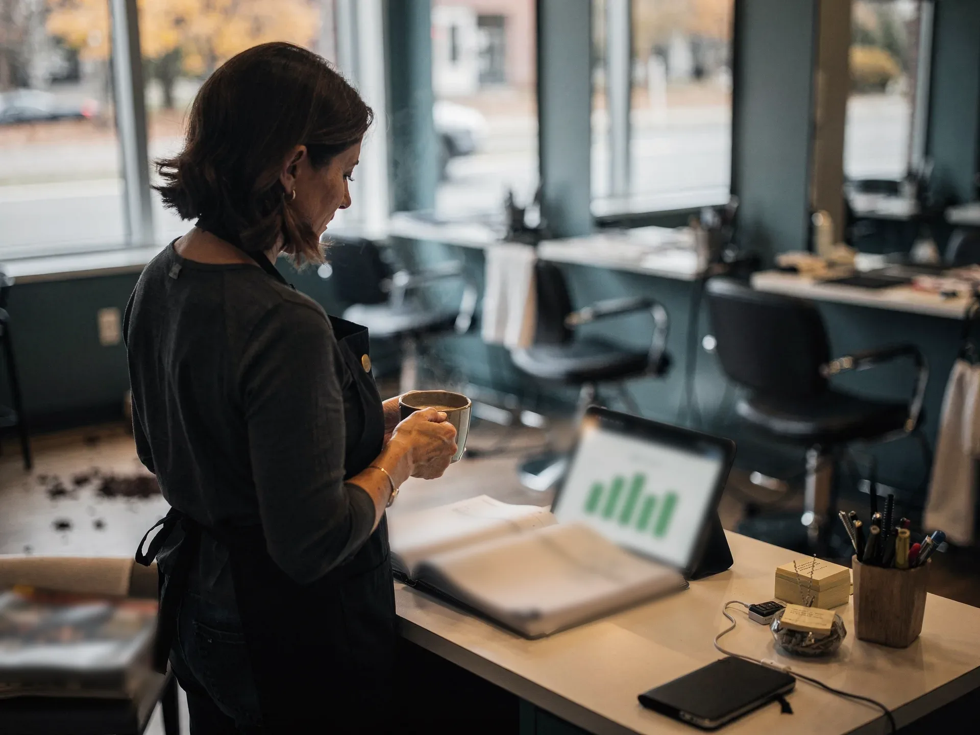 Before opening, a salon owner in a black apron stands at the reception desk with a coffee, reading a green bar chart on a tablet.