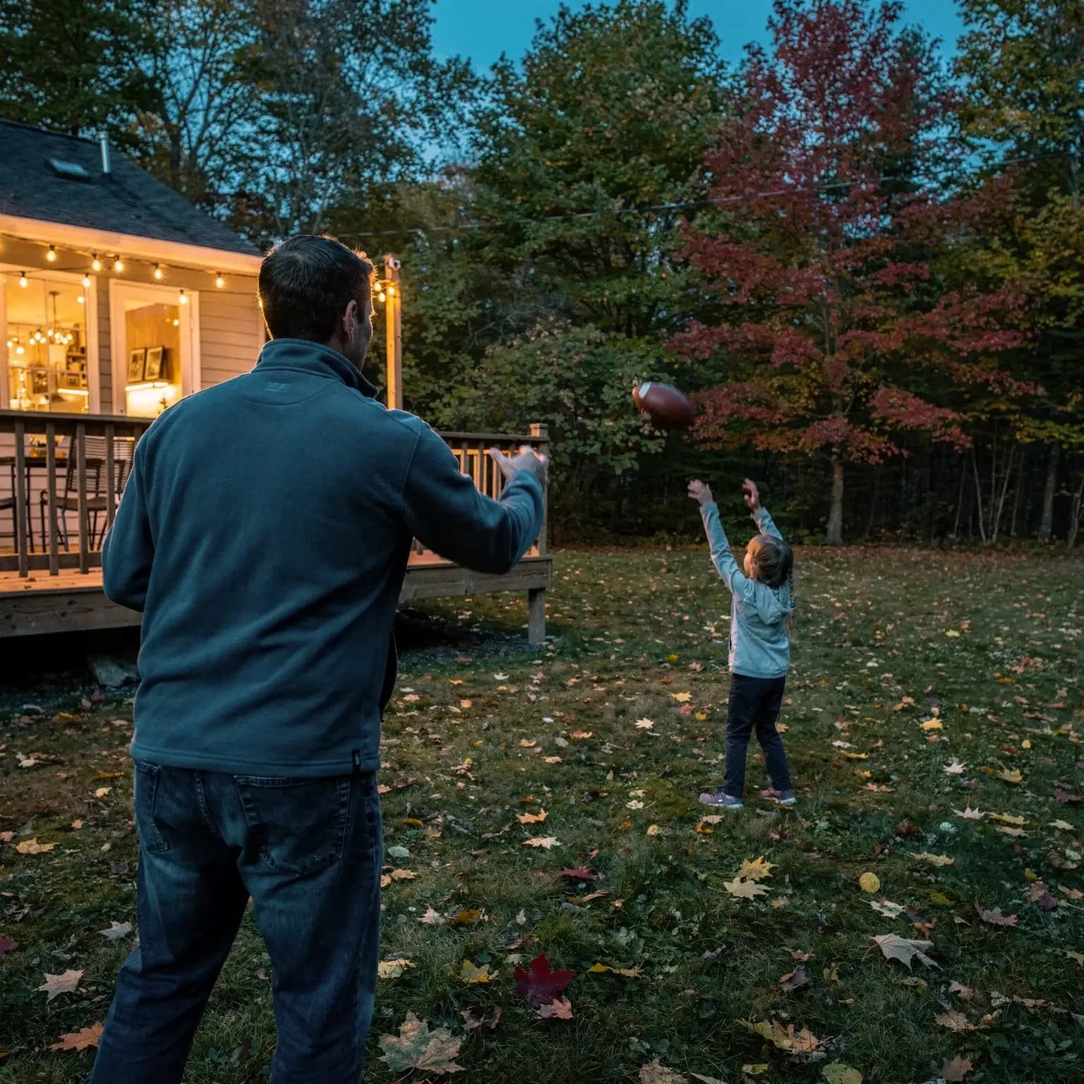 At dusk a father seen from behind tosses a football to his young daughter in a leaf-strewn backyard lit by string lights.