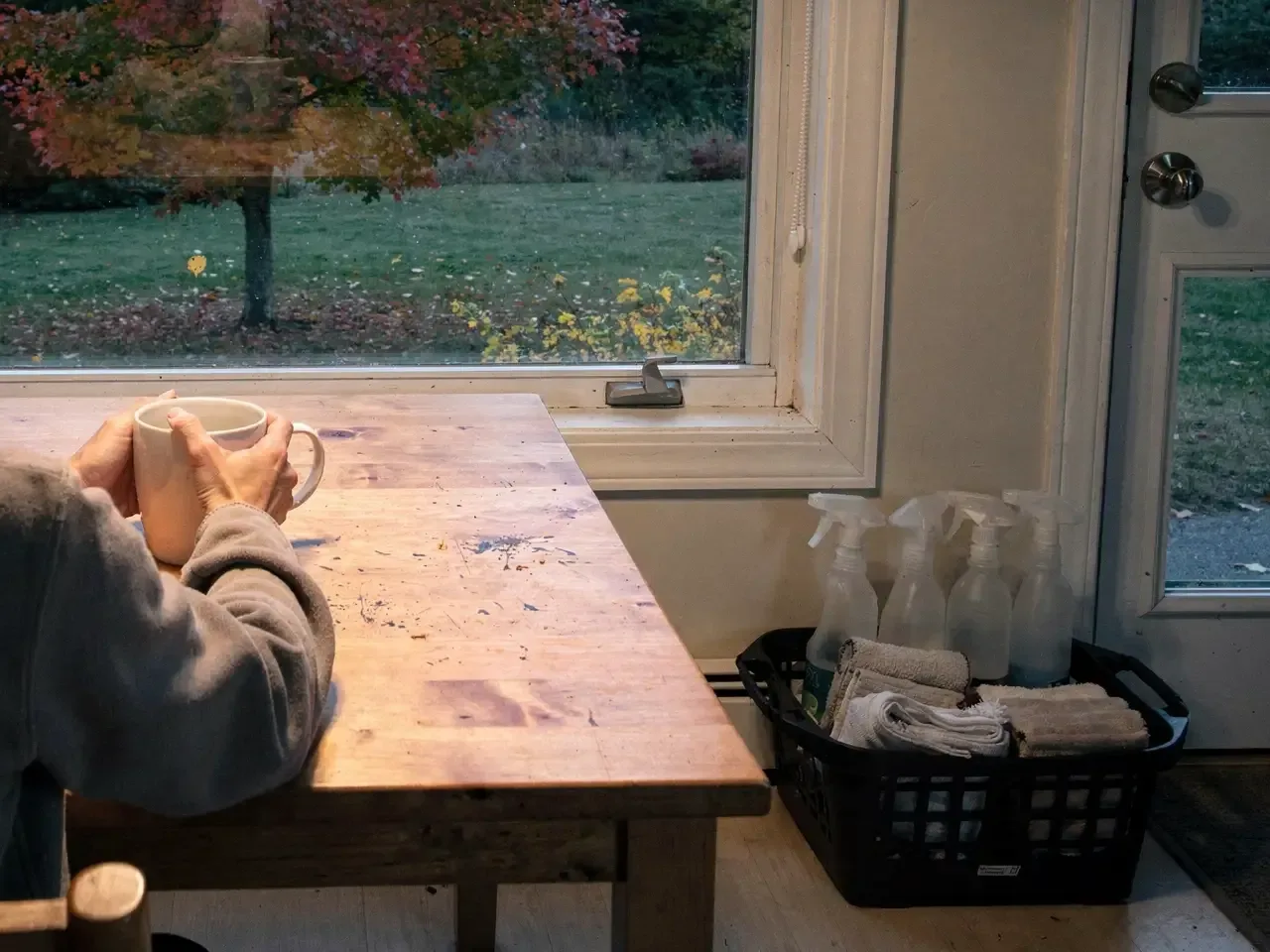 Hands around a mug at a wooden kitchen table at dusk, a basket of spray bottles and folded cloths waiting by the door.