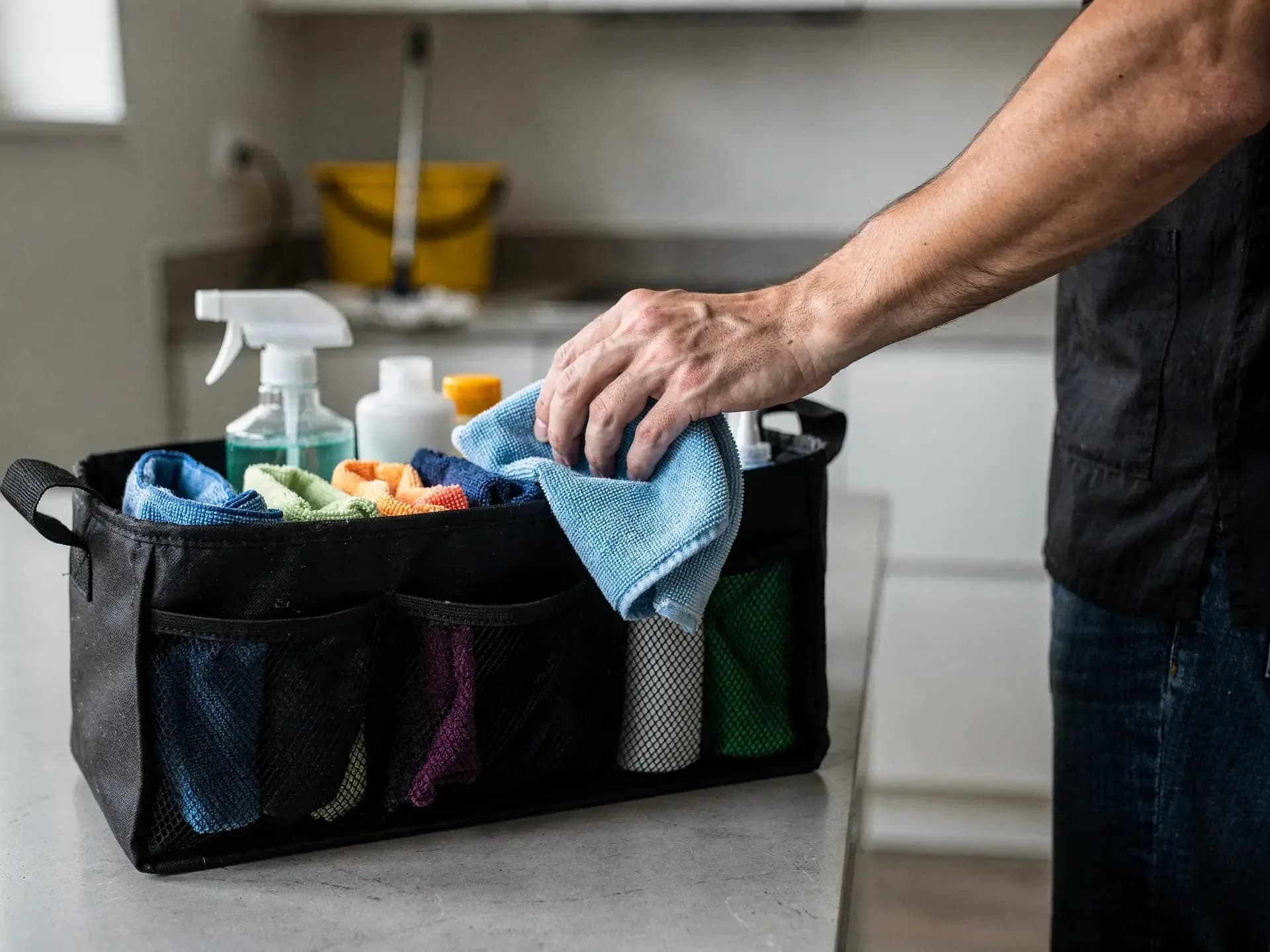 A hand tucks a folded blue cloth into a mesh caddy of color-coded cloths and spray bottles on a kitchen counter.