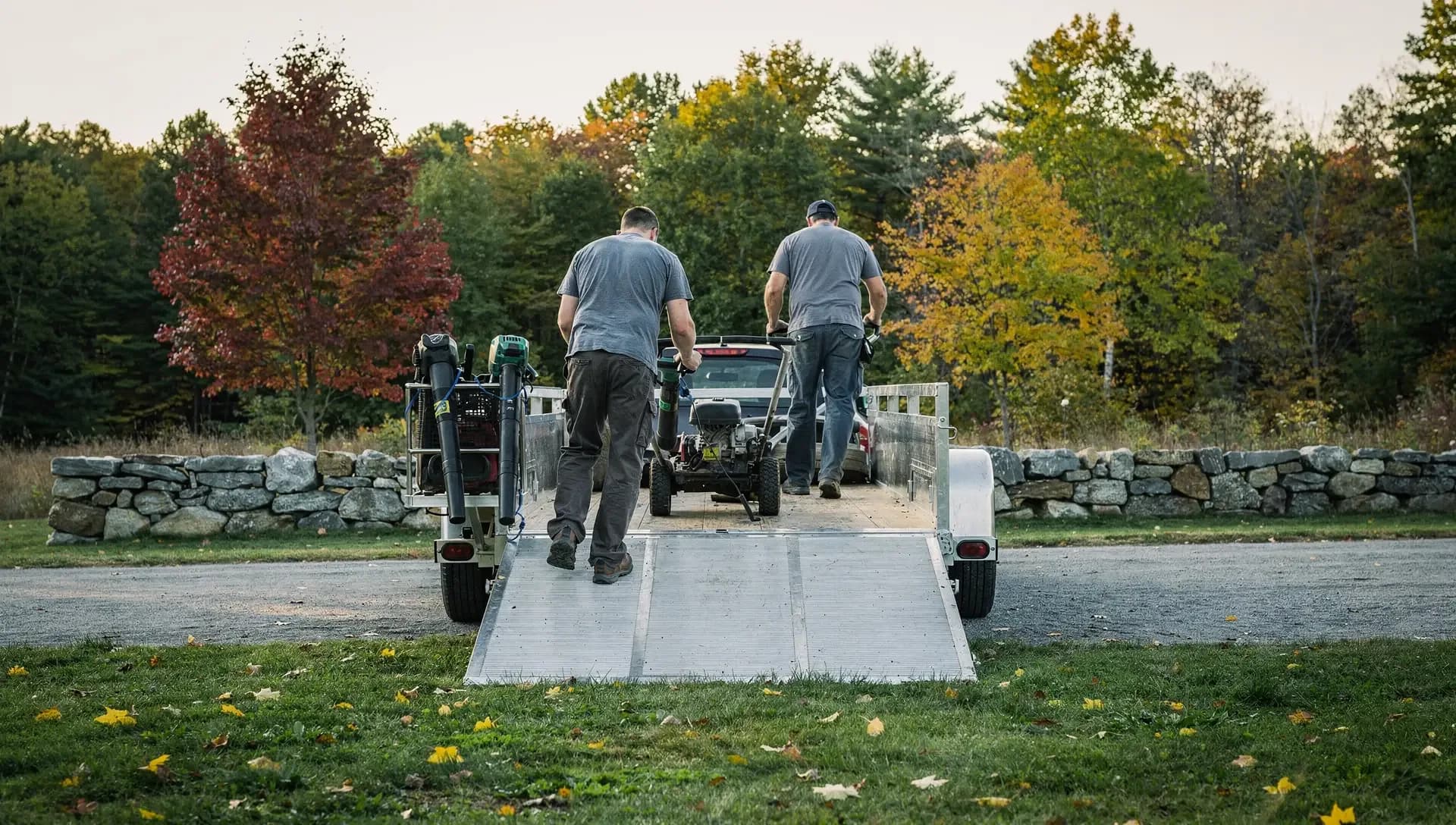 Two crew members wheel walk-behind mowers up the ramp of an open trailer beside a stone wall in early-evening light.