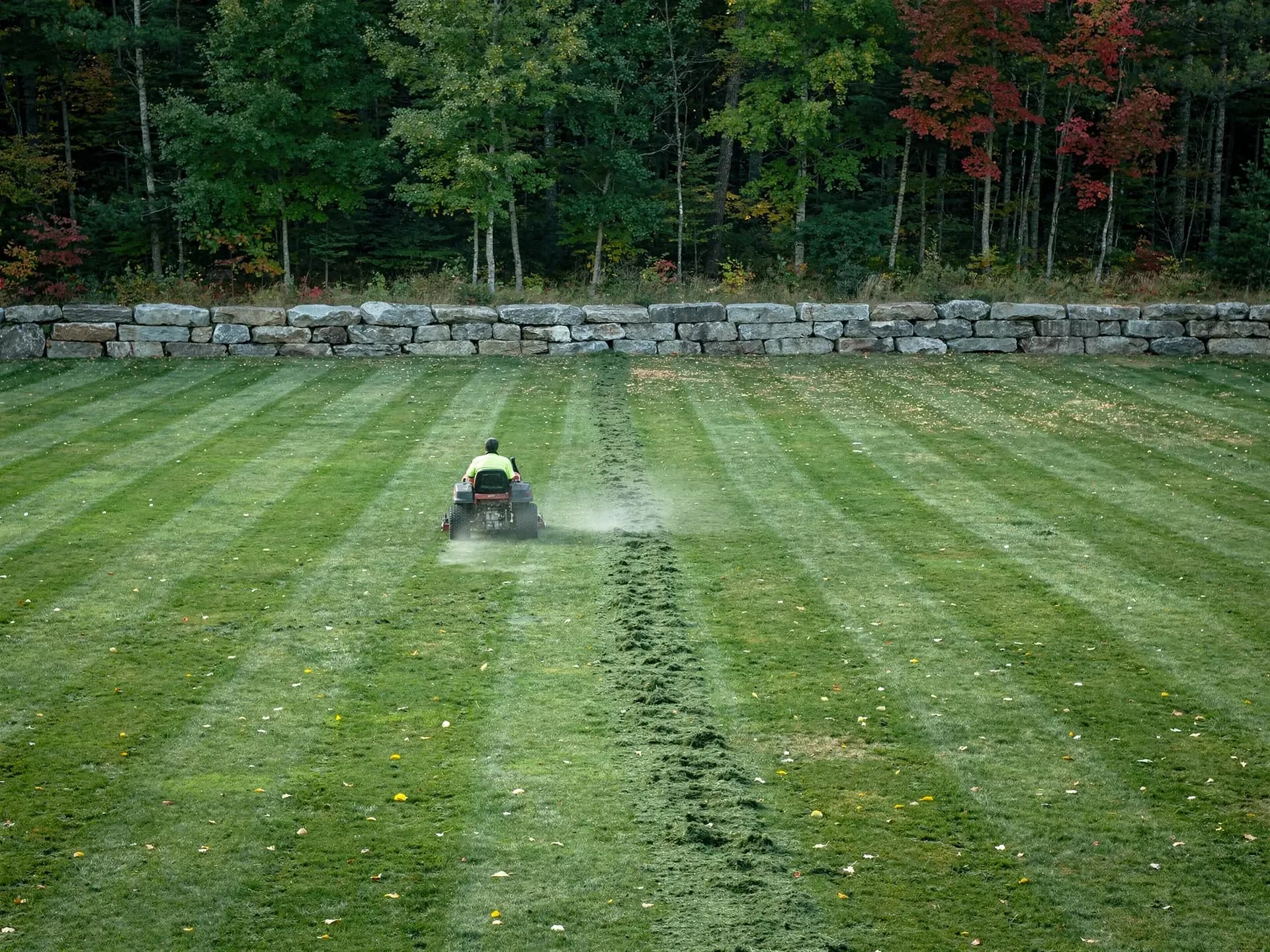 A worker rides a red mower across a freshly striped lawn beside a long granite block wall.