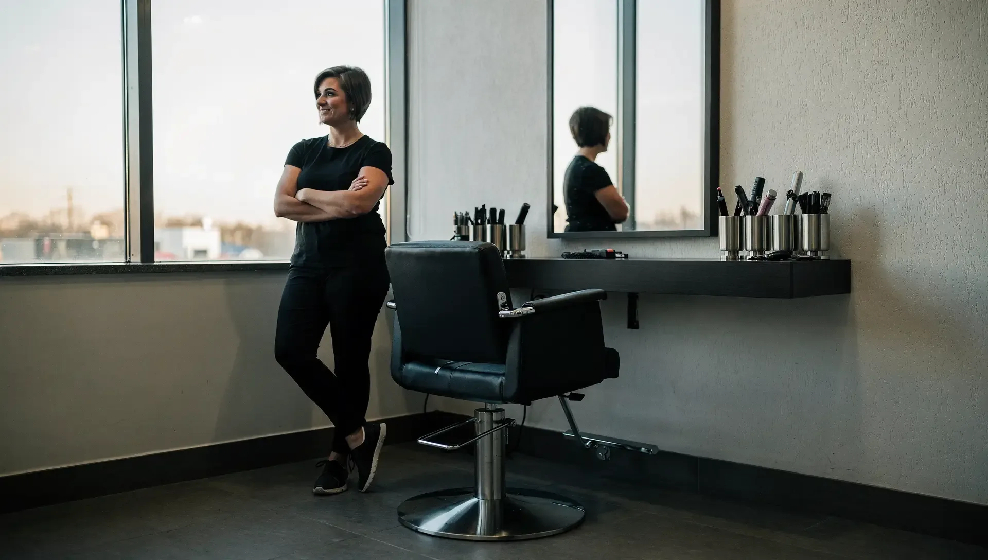 A stylist in black leans by a tall window with arms folded beside an empty salon chair before the day starts.
