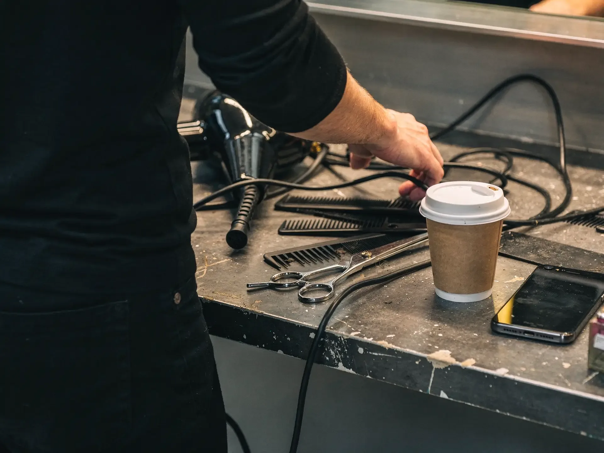 A stylist reaches across a cluttered station tangled with dryer cords, combs, shears and a coffee cup.