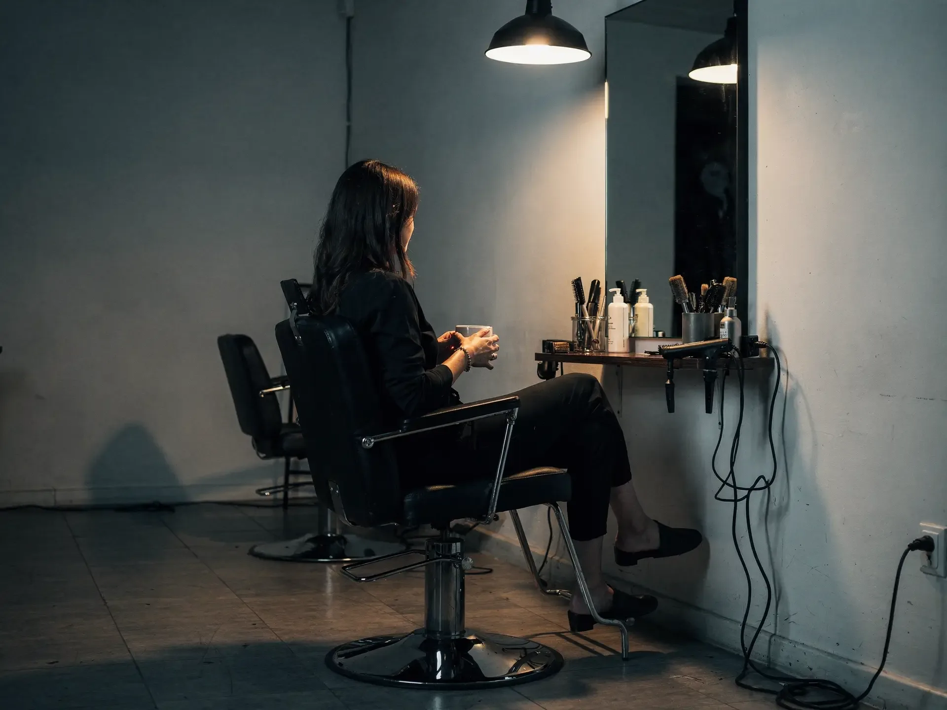 After close, a stylist sits sideways in her own salon chair with a mug under a single pendant lamp.