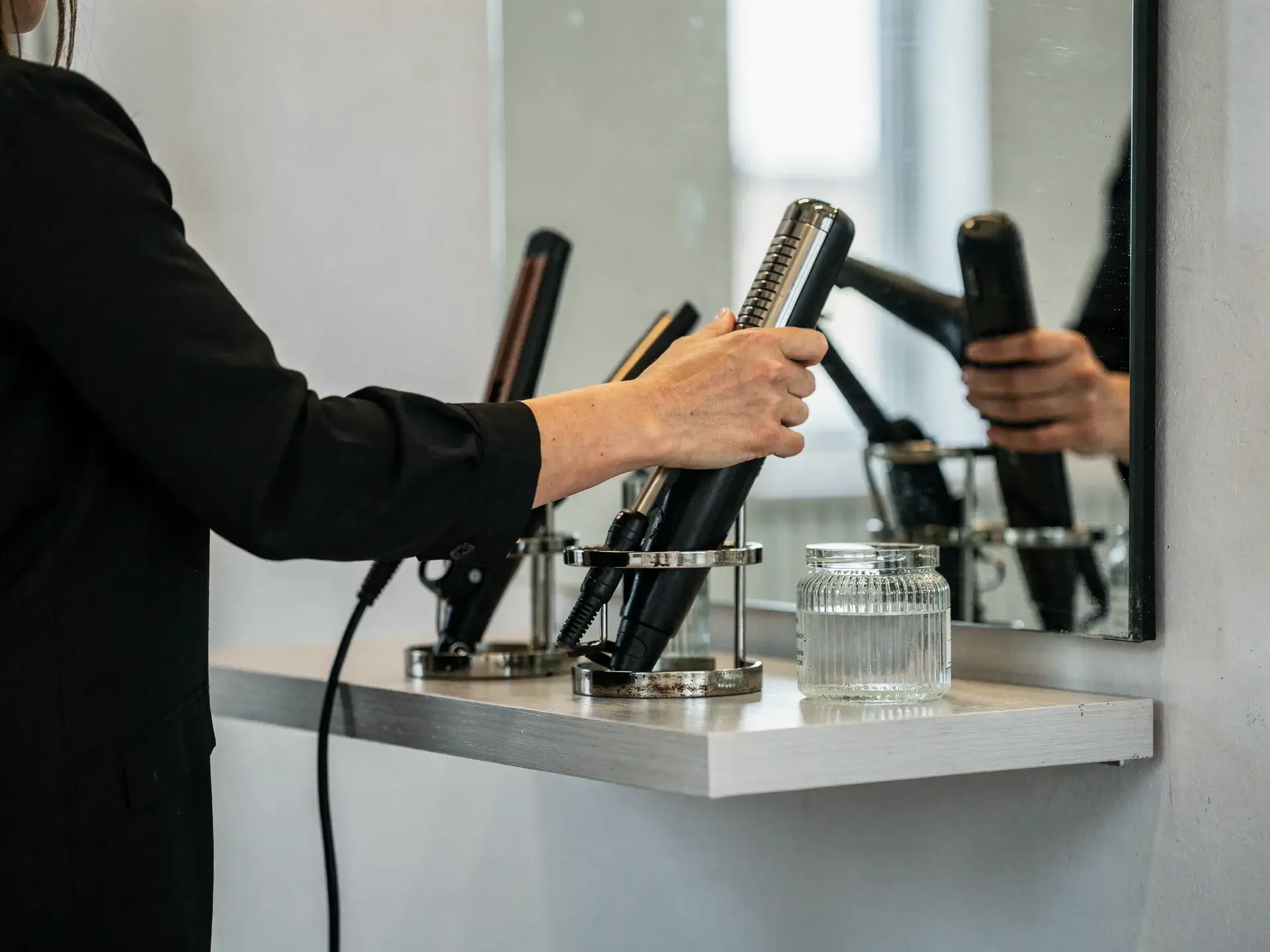 A stylist sets a flat iron into a chrome holder on a pale shelf beside a wall mirror.