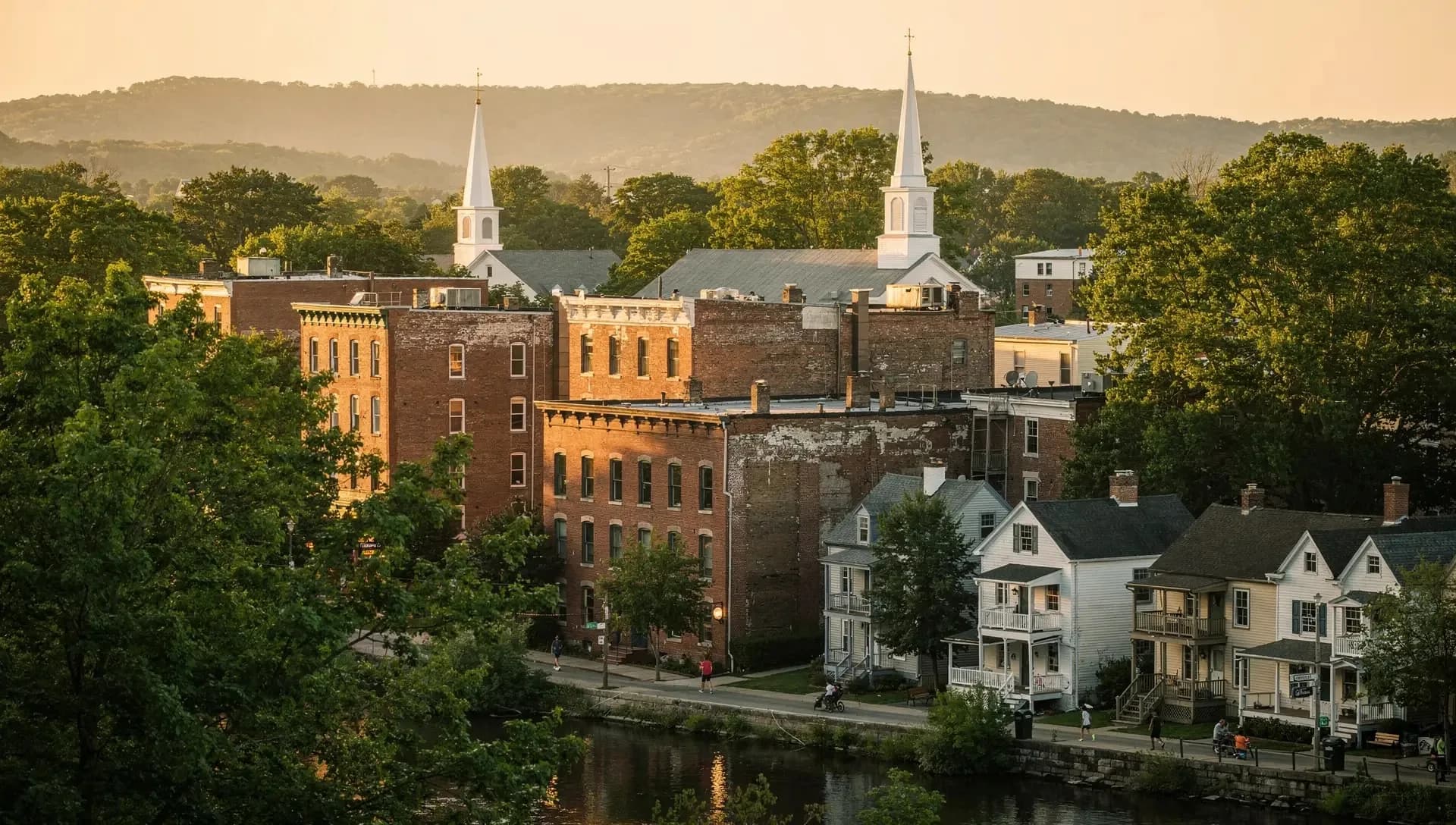 From a hillside at golden hour, red-brick mill blocks and two white church steeples rise above porch-fronted houses along a river, wooded ridges behind.