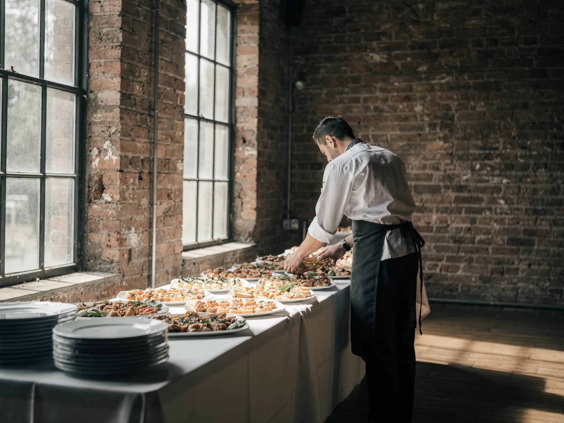 A caterer in a white chef's jacket and dark apron arranges platters along a linen-covered table in a brick-walled room with tall windows.