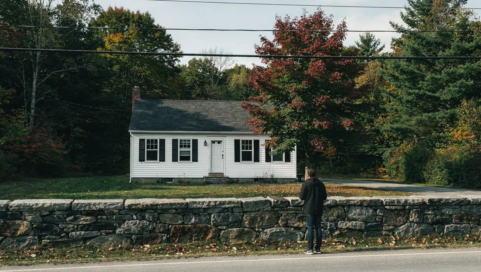 A small white Cape with black shutters sits behind a long fieldstone wall along a two-lane road.