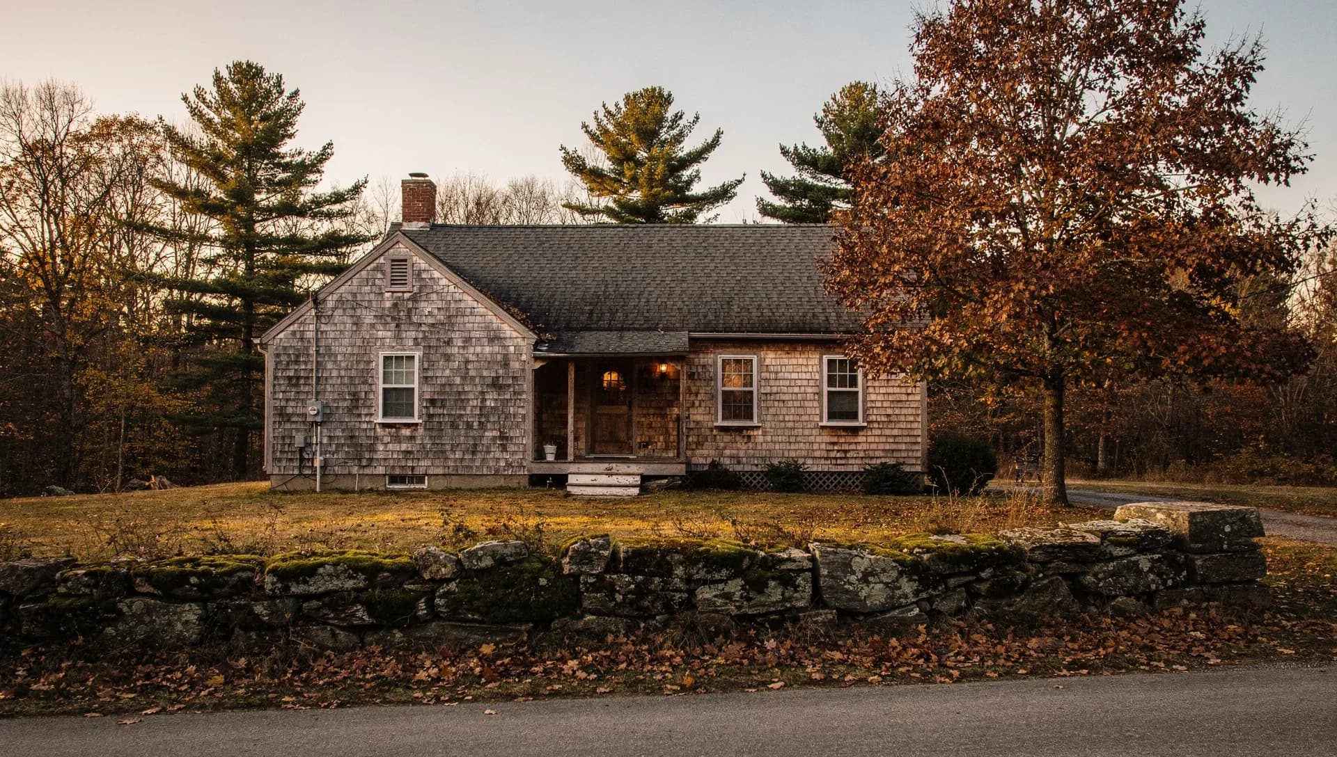 A cedar-shingled Cape with a lit porch sits behind a mossy fieldstone wall at dusk, pines and a russet oak around it.