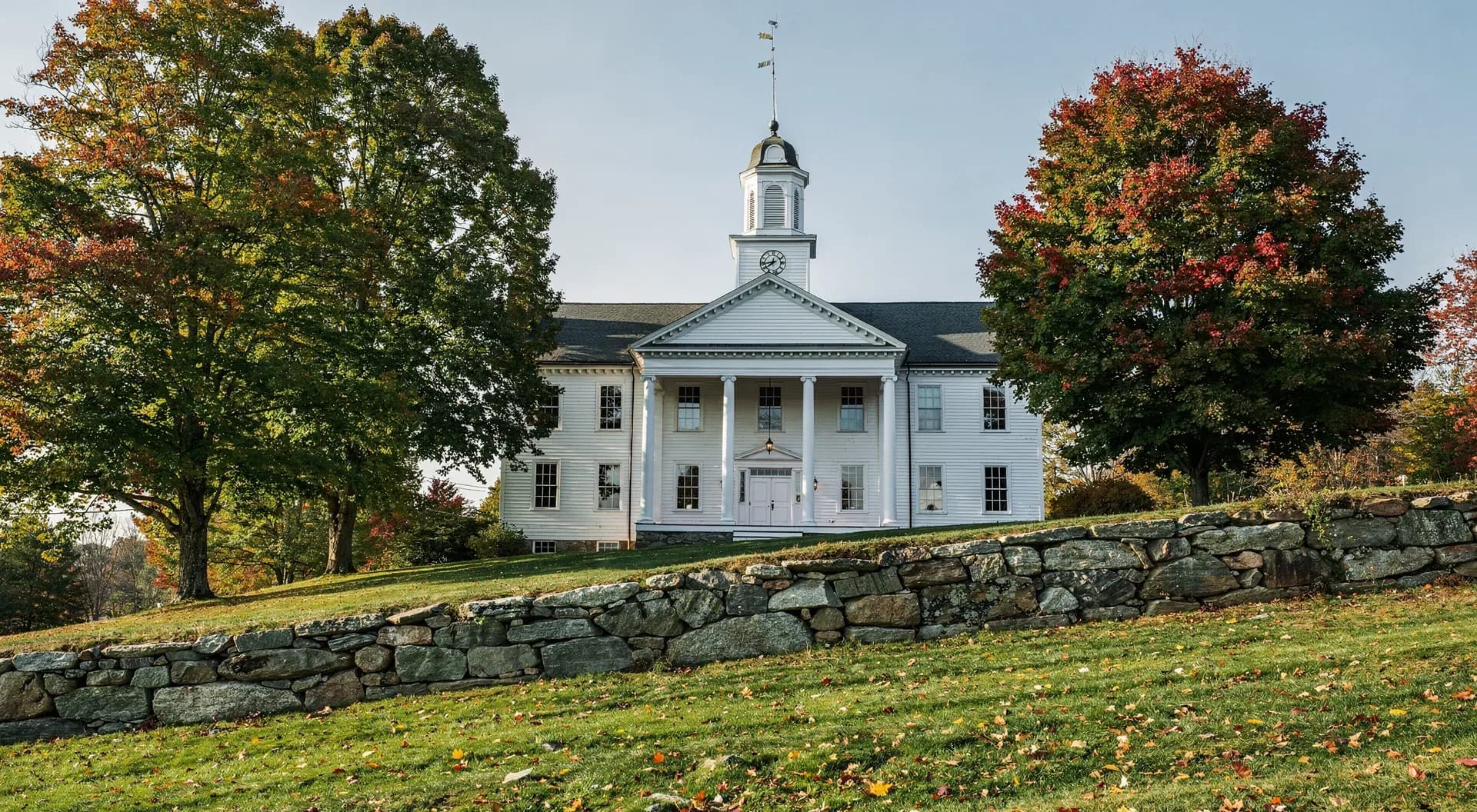 A tall white meetinghouse with a columned porch and a cupola stands on a hill above a fieldstone wall, maples on either side.