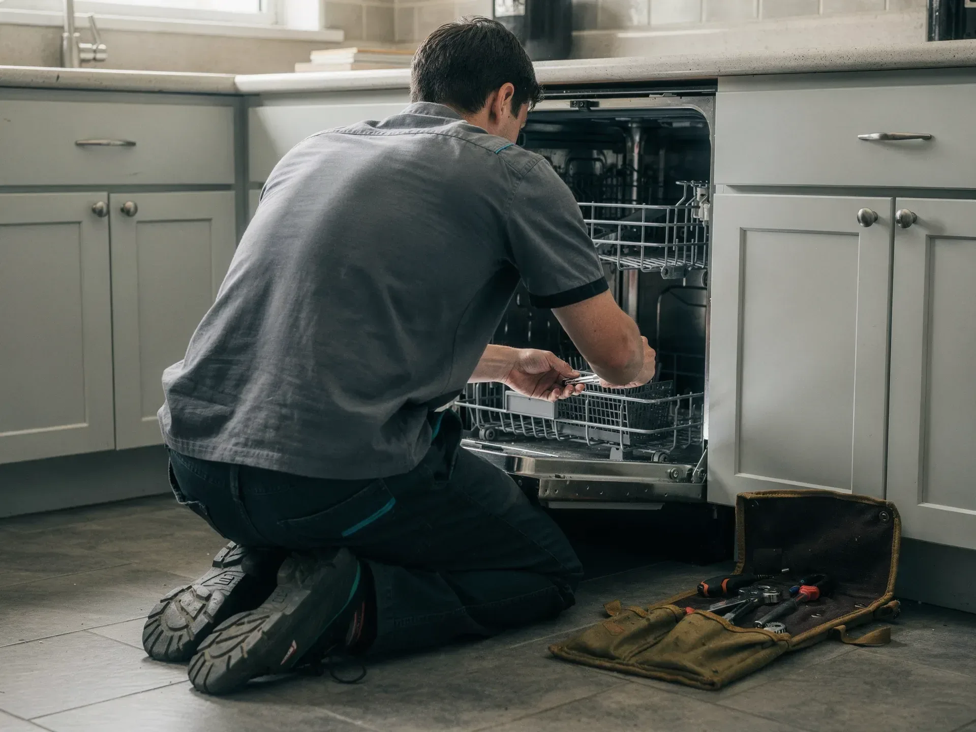 A repair technician in a grey work shirt kneels on a tile kitchen floor reaching into an open dishwasher, a canvas tool roll of screwdrivers and pliers open beside him.