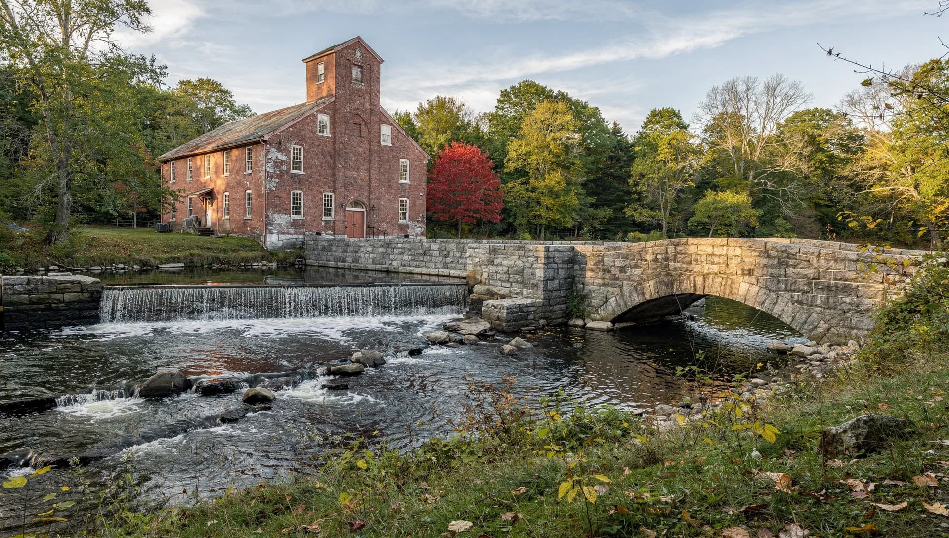 A brick mill with a short tower stands above a stone dam and falls, a stone arch bridge downstream.