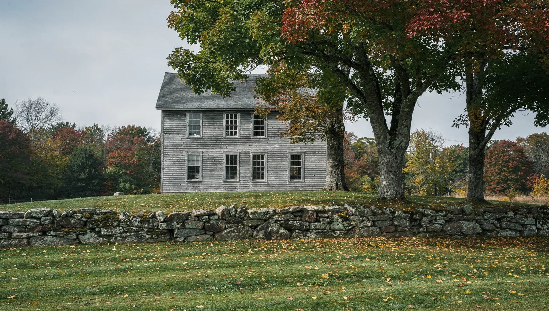 A weathered clapboard meeting house sits on a rise behind a stone wall and two maples.