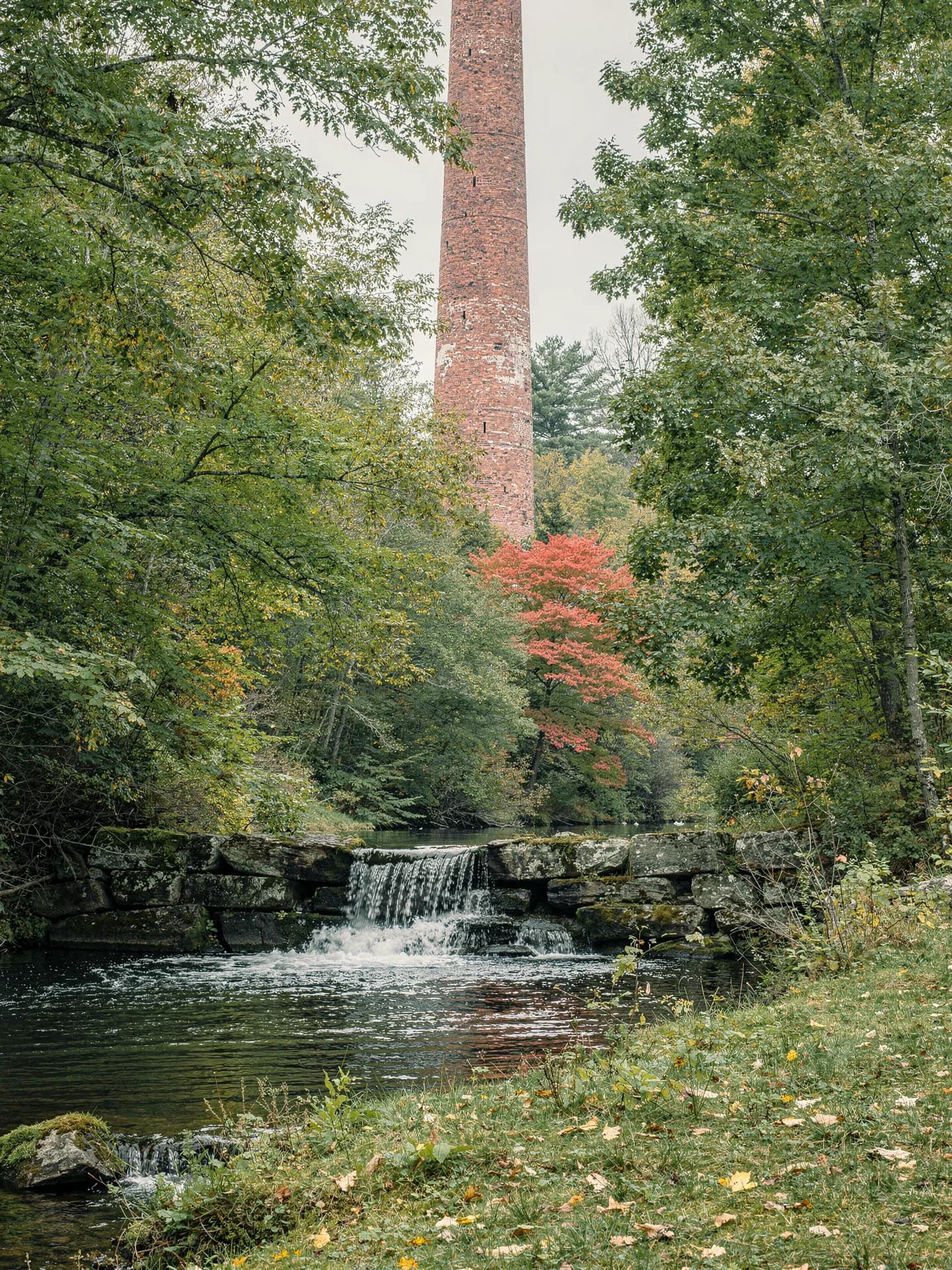 A tall red-brick mill chimney rises through the trees above a brook dropping over a low stone weir.