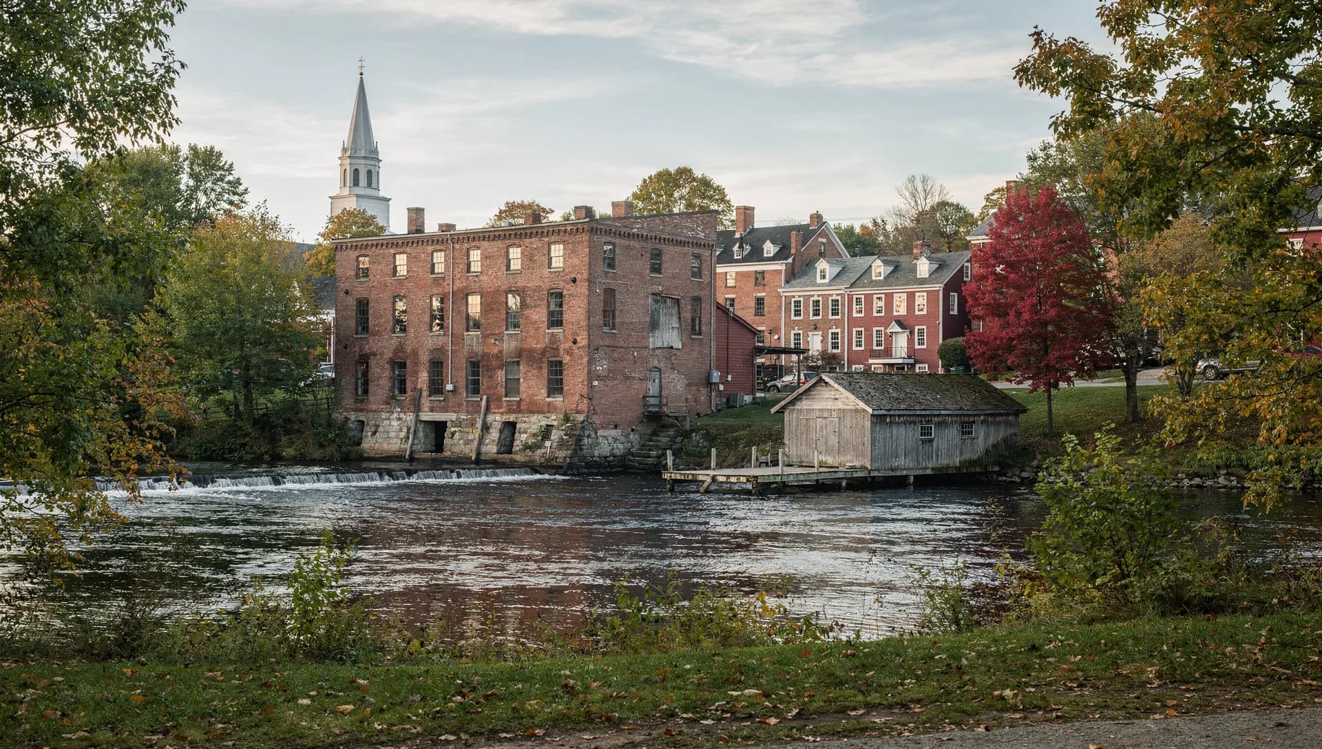 A brick mill stands at a low falls beside a wooden boathouse, a white spire rising over brick rooftops behind.