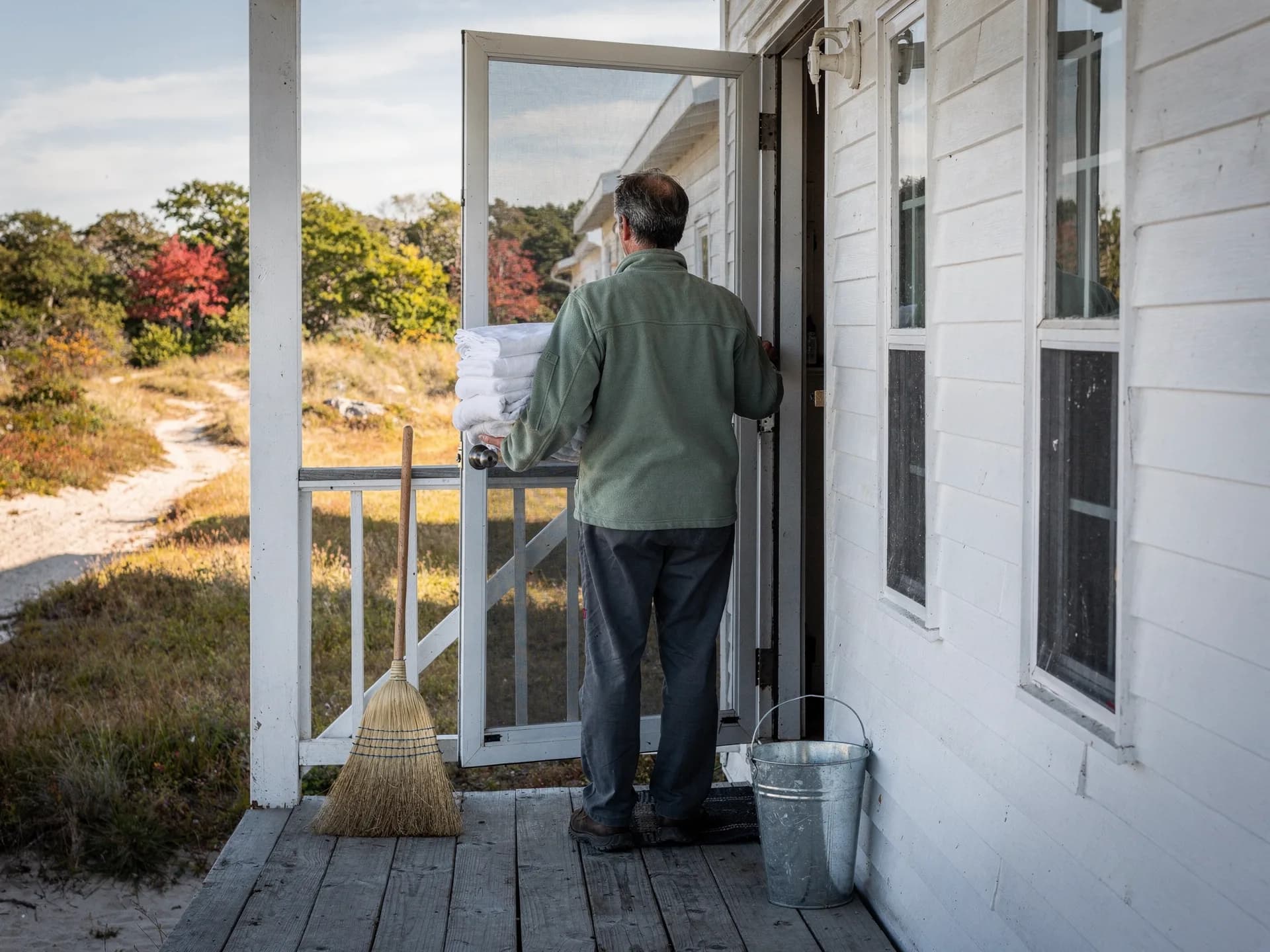 A man in a sage fleece carries a stack of folded white linens through the screen door of a white beach cottage, a broom and a steel bucket on the porch.