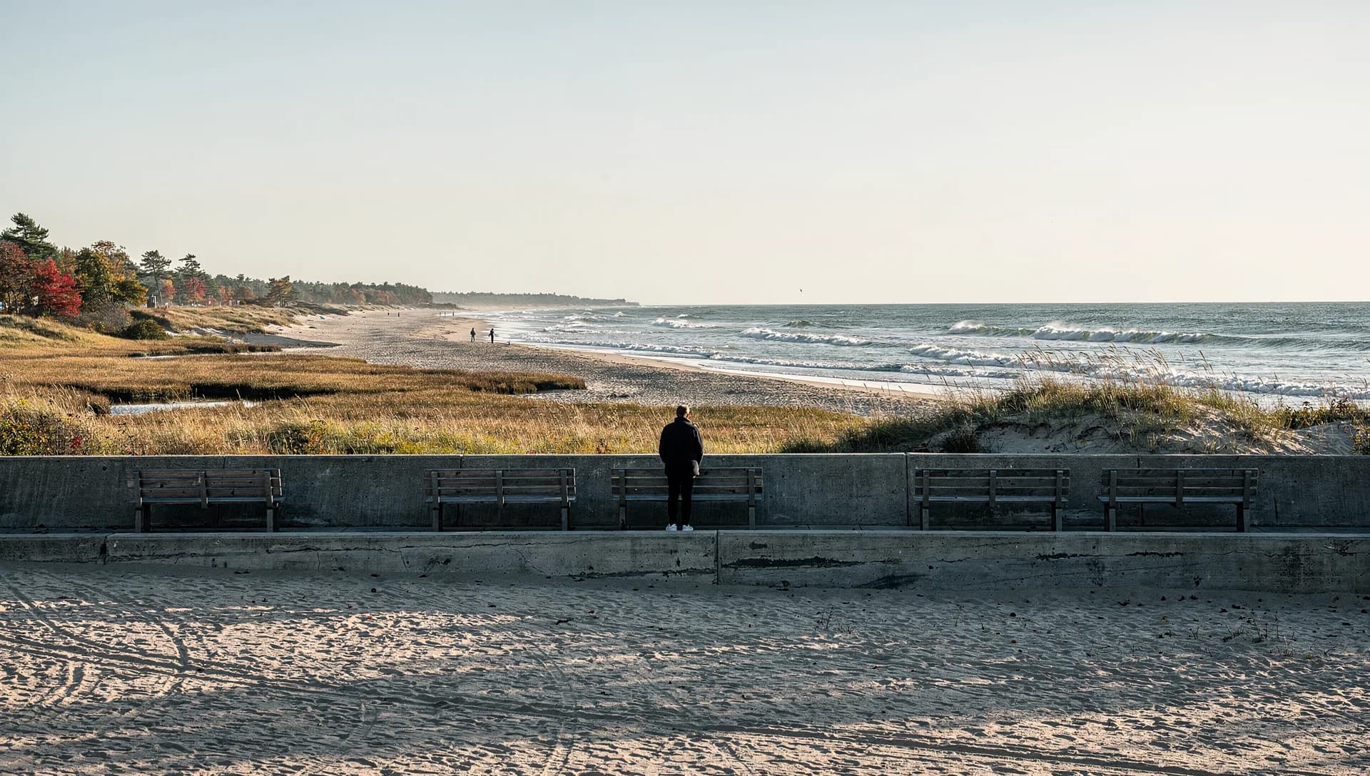 A long empty beach and surf stretch beyond a concrete seawall lined with wooden benches, one person far down the walk.