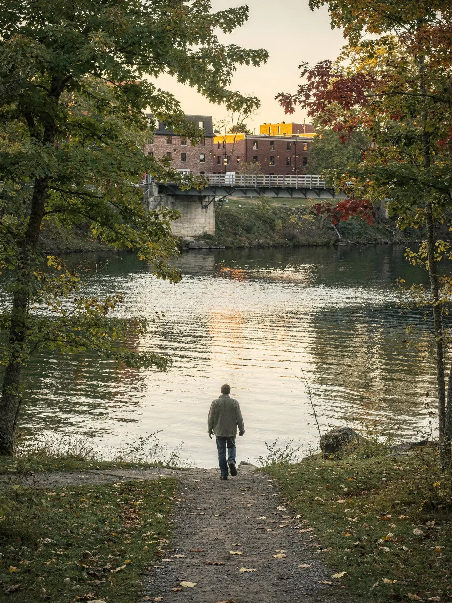 A man walks a dirt path to a wide river, with brick buildings and a bridge in evening light across the water.