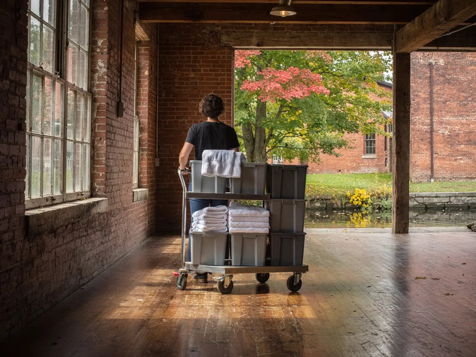 A person pushes a cart of grey bins and white towels across the plank floor of a brick mill toward a canal-side bay.