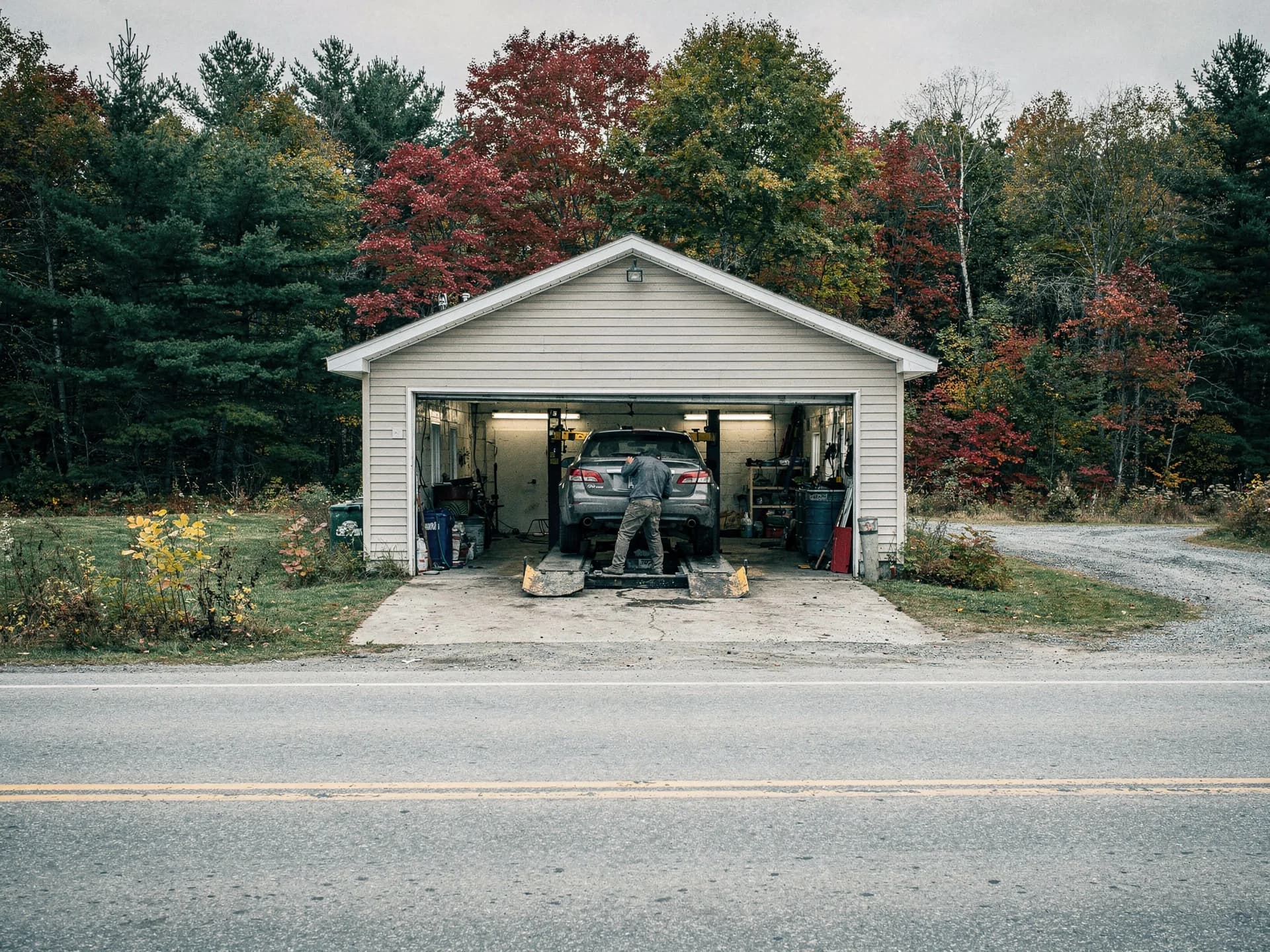 A small garage set back from a two-lane road, its door open on a car up on a lift and a mechanic at work.