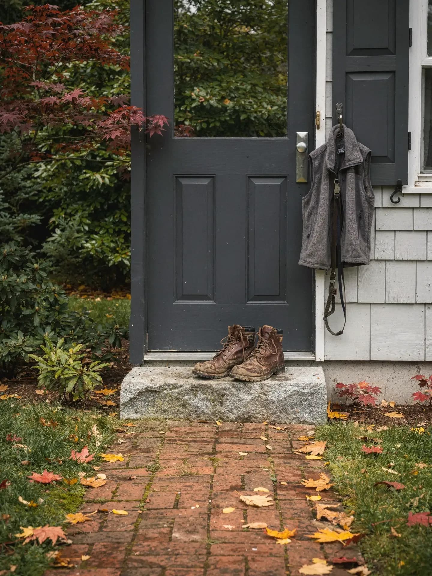 Muddy work boots sit on a granite step by a grey door, a fleece vest and a dog leash hanging on a hook.