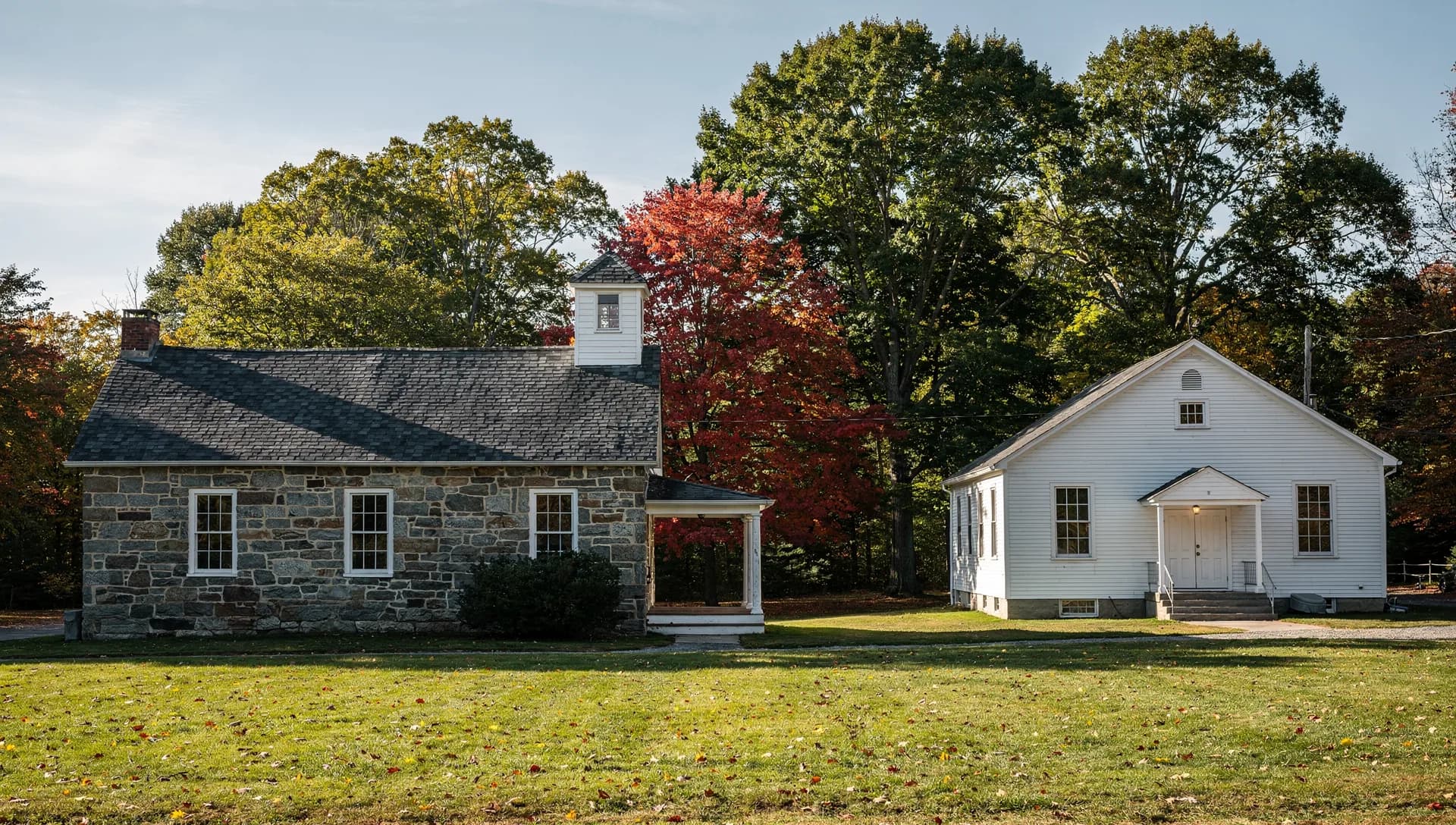 A fieldstone building with a slate roof and a small cupola stands beside a white clapboard hall, maples behind a mown green.