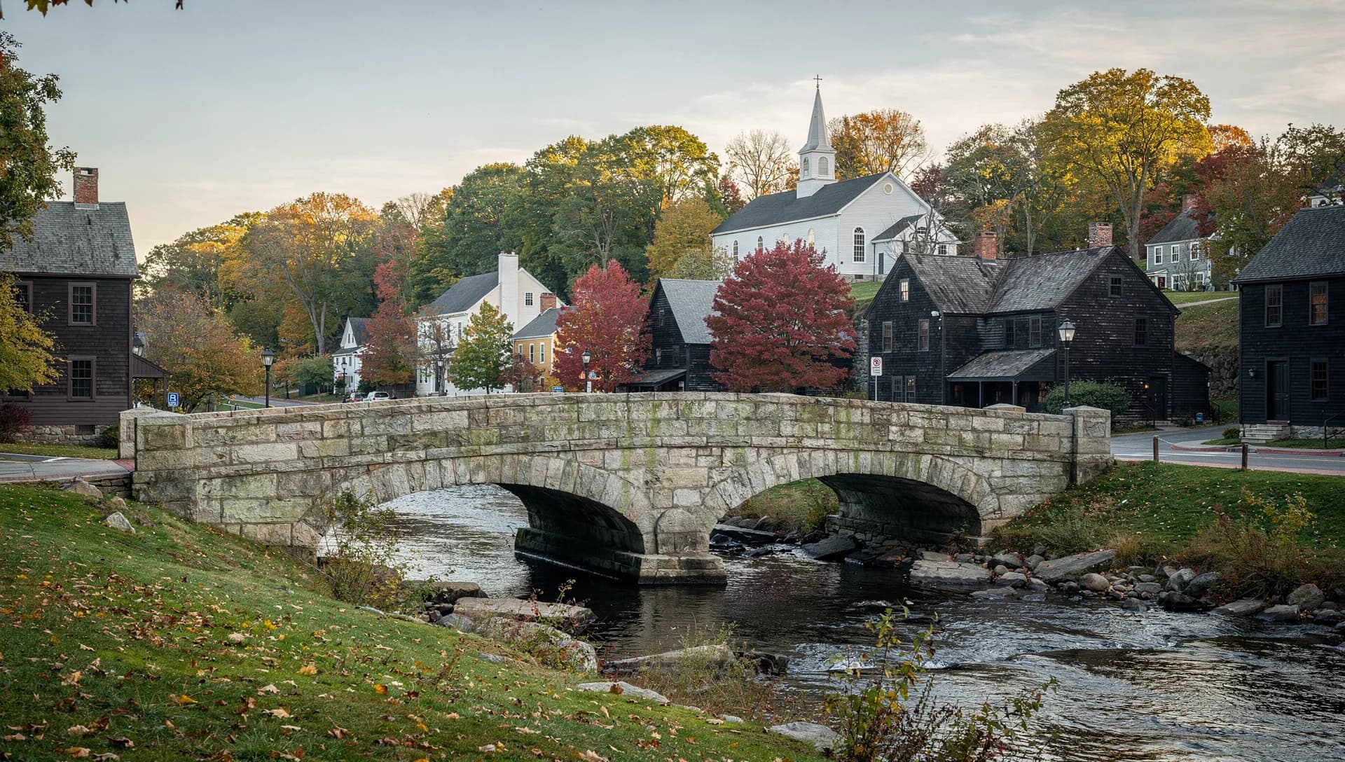 A double-arched stone bridge crosses a small river below dark old timber houses and a white church.