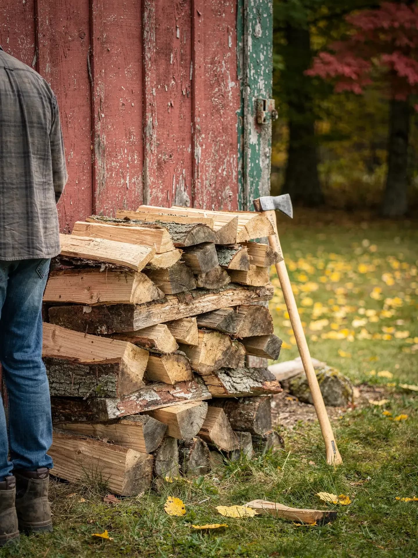 A man stands beside a stack of fresh-split firewood against a peeling red barn door, an axe leaning nearby.