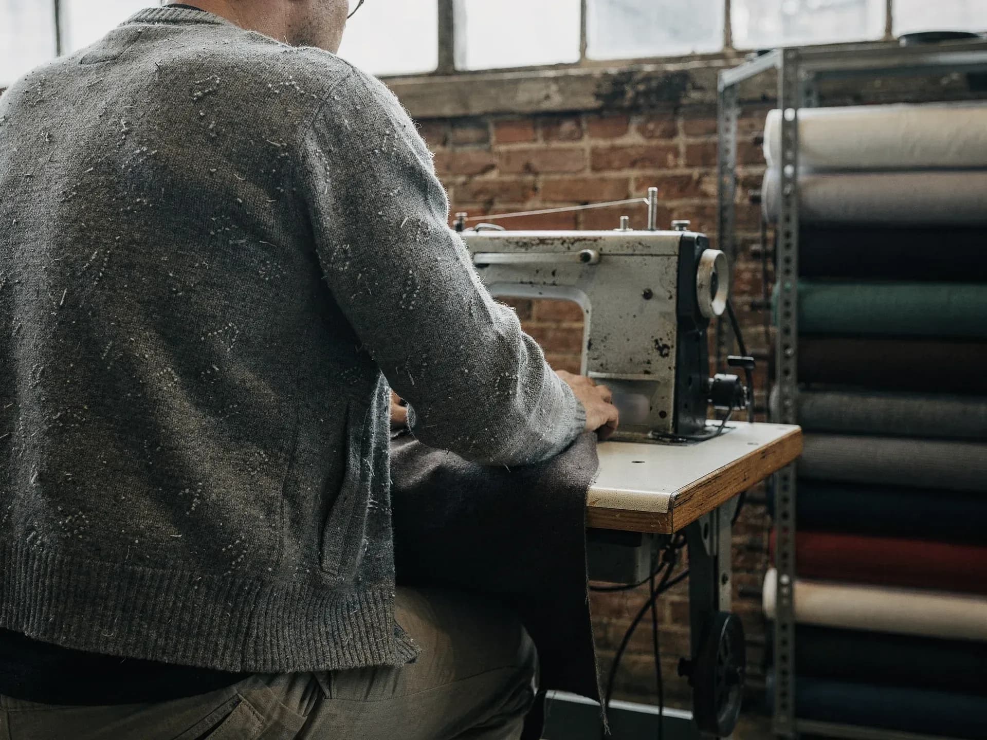 A tailor in a grey cardigan feeds dark cloth through an old industrial sewing machine, bolts of fabric shelved against a brick wall.