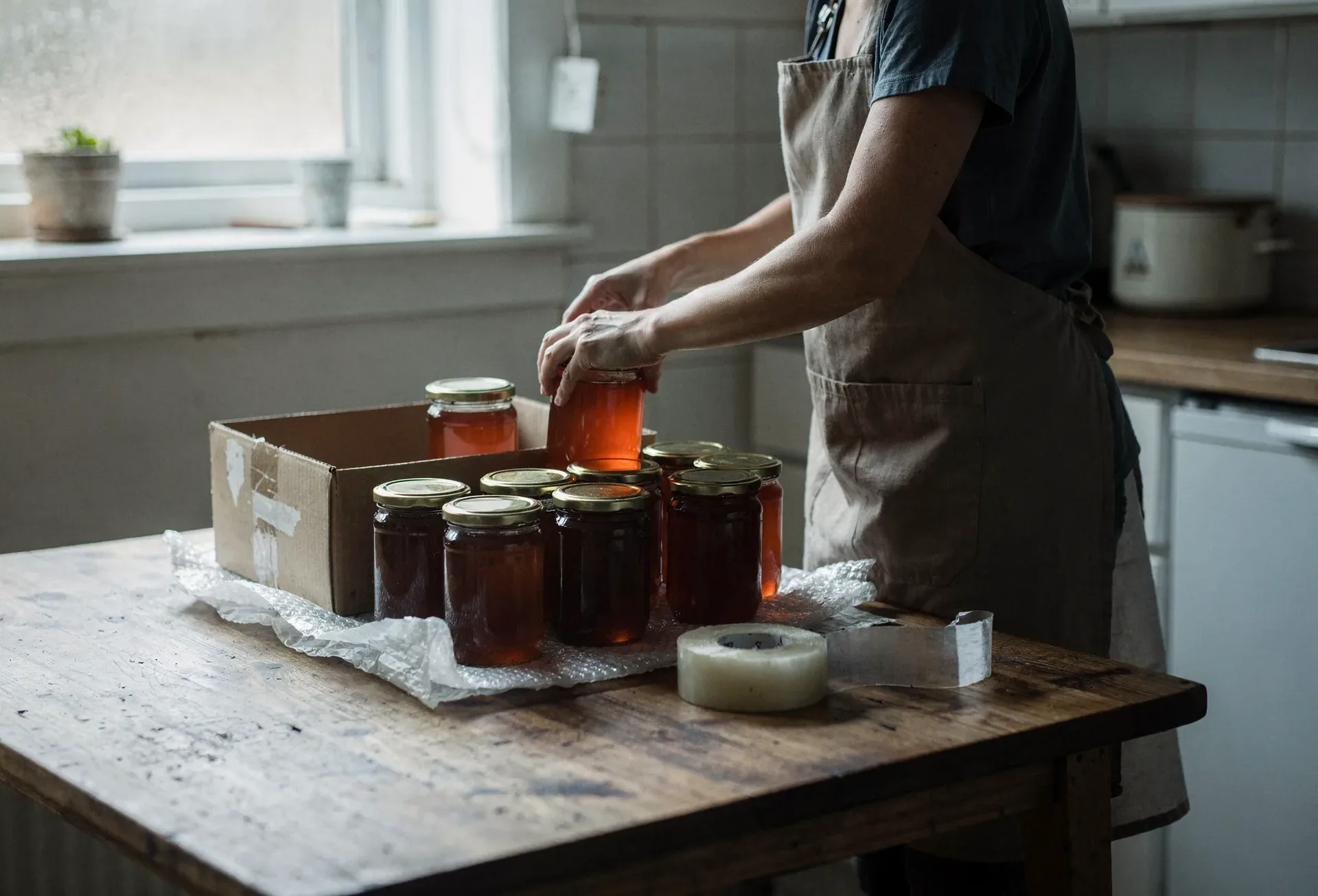 A woman in a linen apron packs jars of amber honey into a cardboard box at a wooden kitchen table, a roll of tape beside her.
