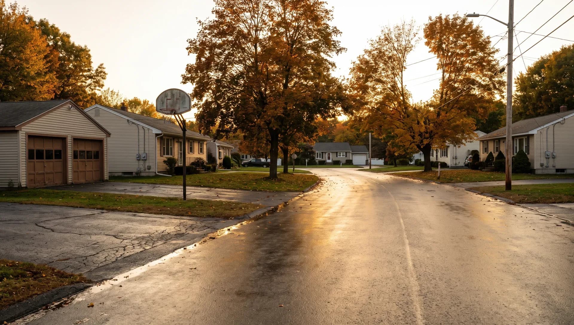 A rain-wet street curves past ranch houses and a two-door garage under autumn maples, a basketball hoop beside one driveway.