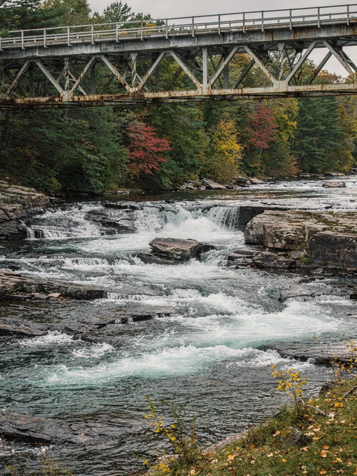 A river tumbles over ledges of dark rock below a steel truss bridge, autumn trees on the far bank.